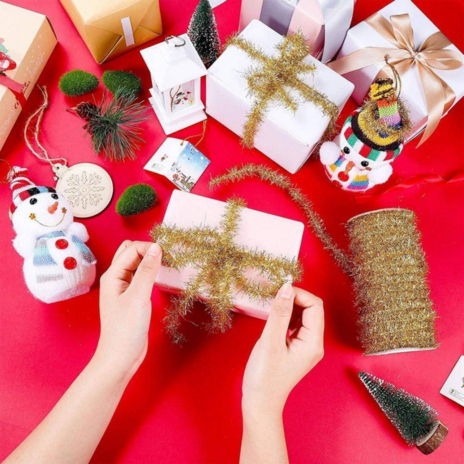 Hände wickeln eine weiße Geschenkbox mit goldenem Lamettaband auf einer roten Oberfläche ein, umgeben von anderen dekorierten Geschenken und kleinen Weihnachtskugeln.