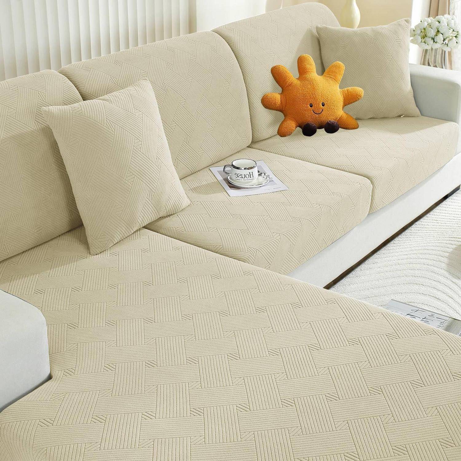 Beiges Ecksofa mit strukturierter Bezug, zwei passende Kissen, ein Tablett mit Tasse und Zeitschrift, und ein orangefarbiges, sonnenförmiges Plüschkissen in einem hellen Wohnzimmer.