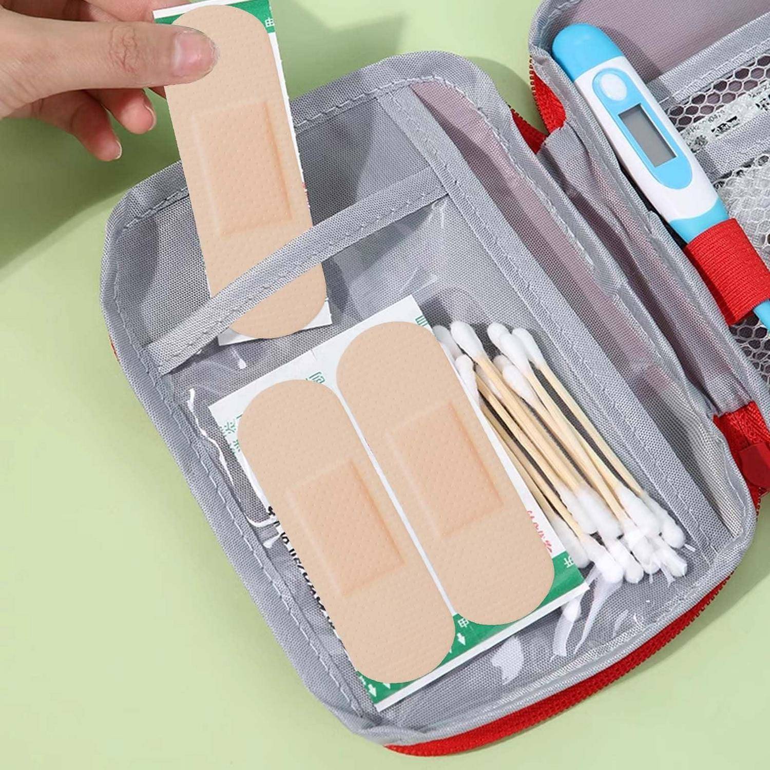 Erste-Hilfe-Set mit Verbandsmaterialien, Wattestäbchen und einem Thermometer in einer grauen Tasche mit Reißverschluss; eine Hand hält ein Pflaster darüber.