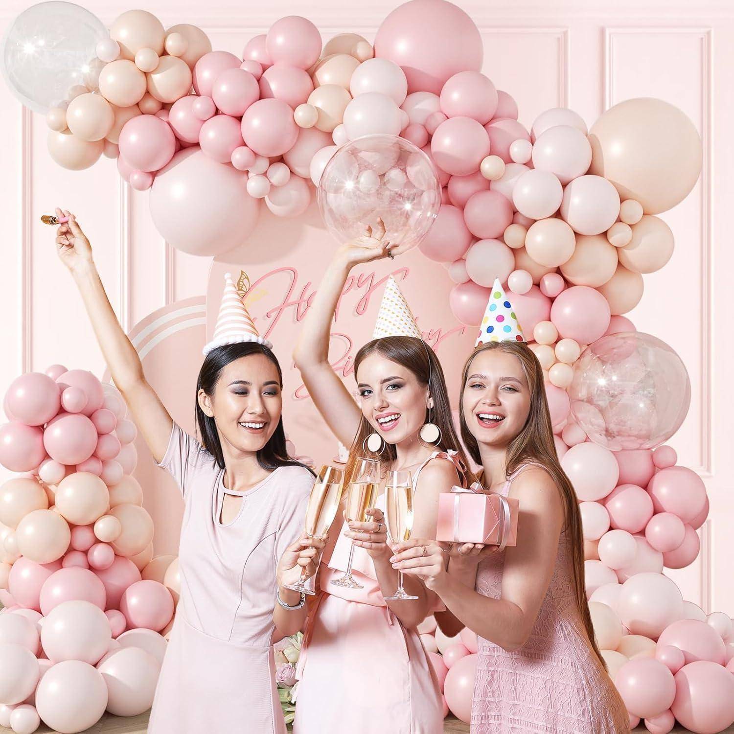 Drei Frauen in rosa Partykleidern und Hüten feiern vor einem rosa Luftballonhintergrund, halten Champagner und eine Geschenkbox.