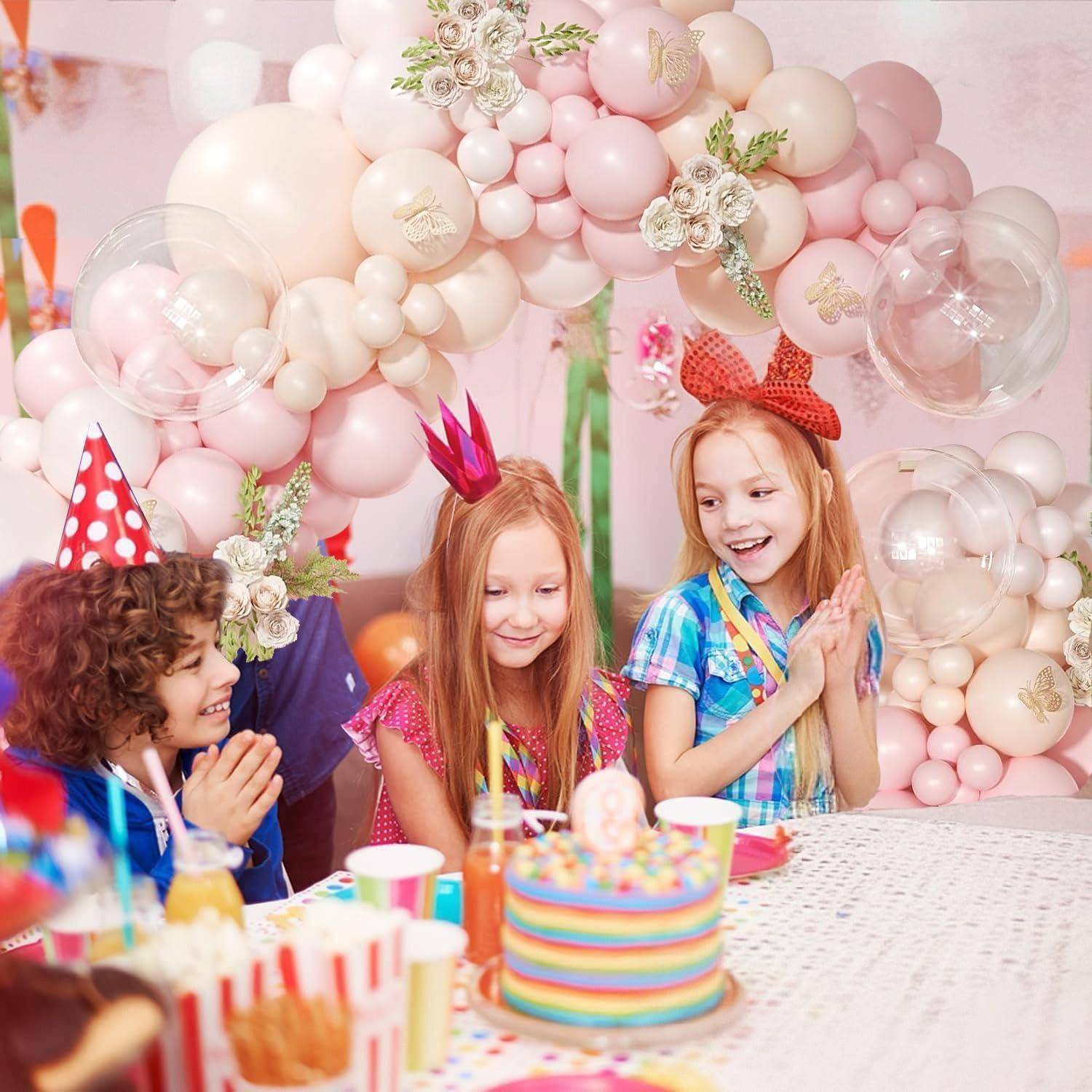 Drei Kinder in Partyhüten feiern an einem Tisch mit Pastellballondekoration zu einem Regenbogenkuchen und Snacks.