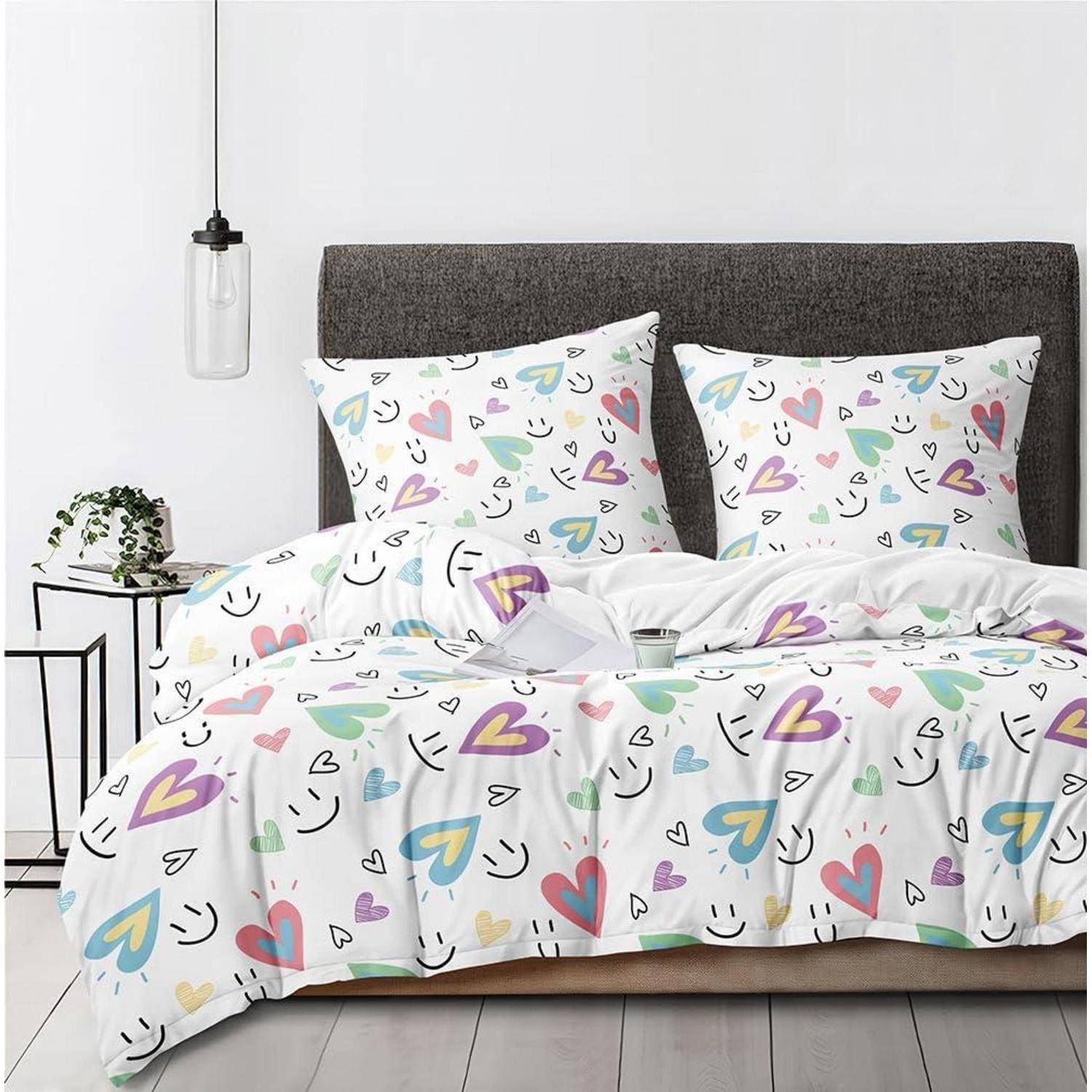 Buntes herzgemustertes Bettzug auf einem ordentlich gemachten Bett mit grauem Kopfteil, weißen Wänden und einem kleinen Nachttisch mit einer Pflanze und einer Pendelleuchte.