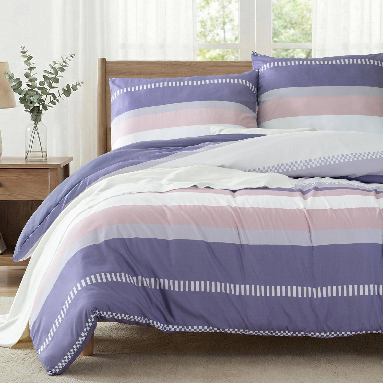 Ein ordentlich gemachtes Bett mit violett, rosa und weiß gestreifter Bettwäsche und Kissen in einem hellen Schlafzimmer neben einem hölzernen Nachttisch und einer Vase mit Grünpflanzen.