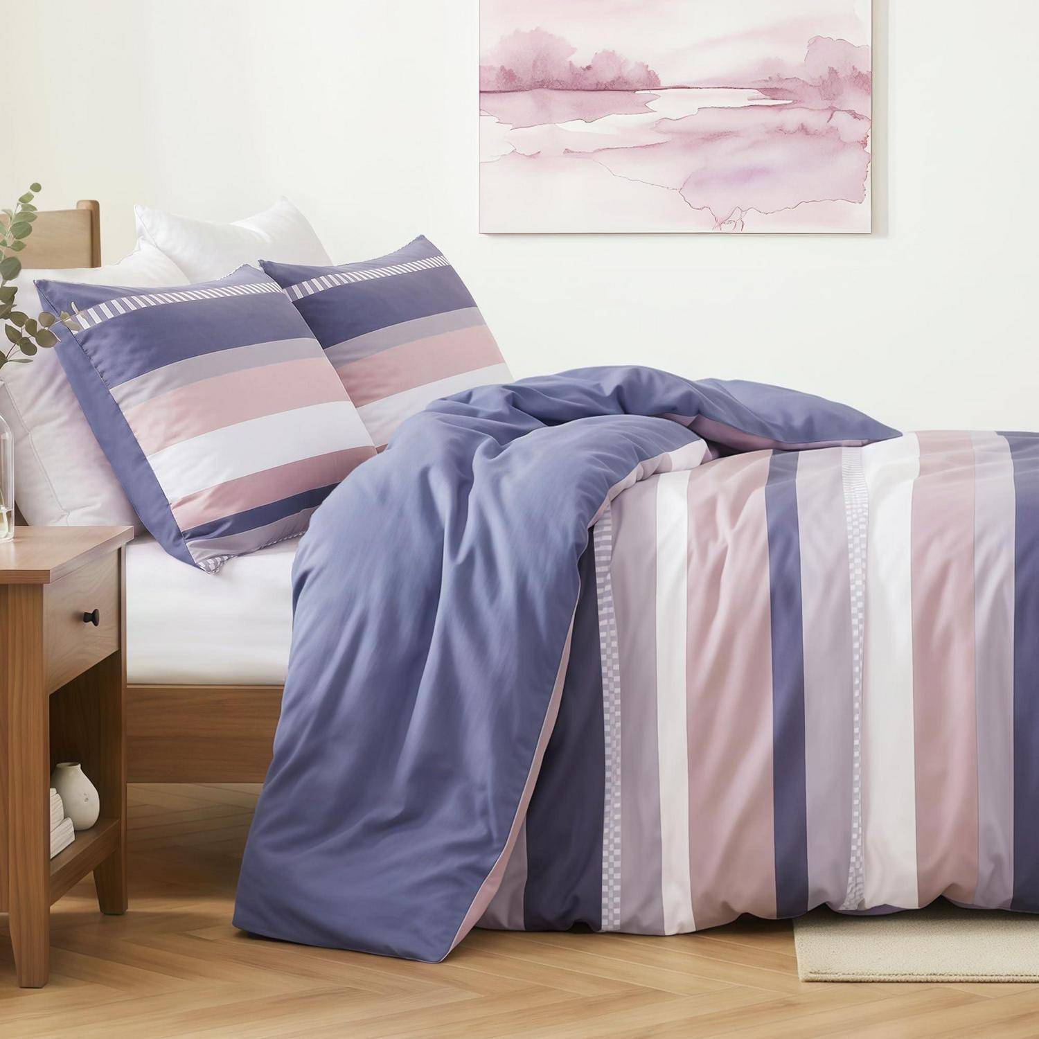 Ordentlich gemachtes Bett mit violett-, rosa- und weiß gestreifter Bettwäsche und Kissen in einem hellen Schlafzimmer neben einem hölzernen Nachttisch und pastellfarbener Wandkunst.