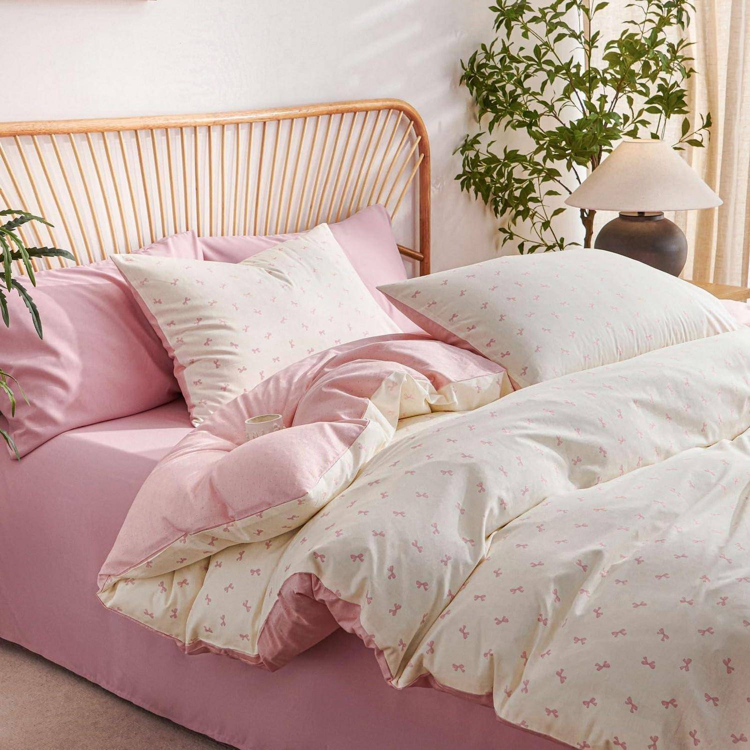 Rosa Bettwäsche mit einer cremefarbenen Blumen-Steppdecke und Kissen auf einem Holzbett, stilisiert in einem hellen Schlafzimmer mit Pflanzen und einer Nachttischlampe.