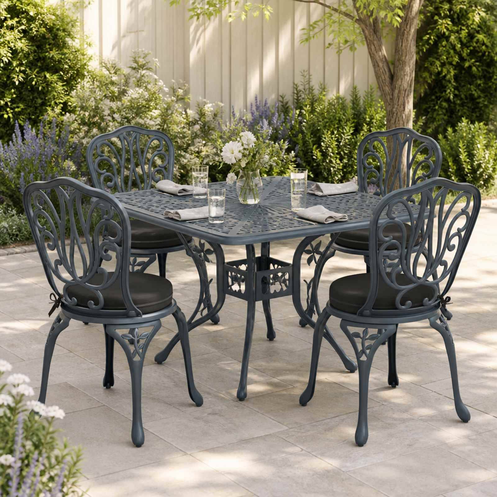 Vier verzierte schwarze Metallterrassen­stühle um einen quadratischen Außenesstisch mit Glastischplatte, eingedeckt mit Gläsern, Servietten und einem Blumen­arrangement als Tischzentrum in einem Garten.