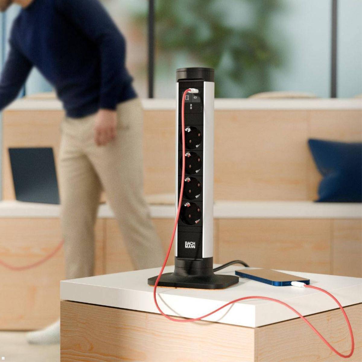 Schwarze Steckdosenleiste auf einem Tisch in einem Loungebereich mit vier Steckdosen und roten/schwarzen Kabeln, mit einer unscharfen Person, die dahinter vorbeigeht.