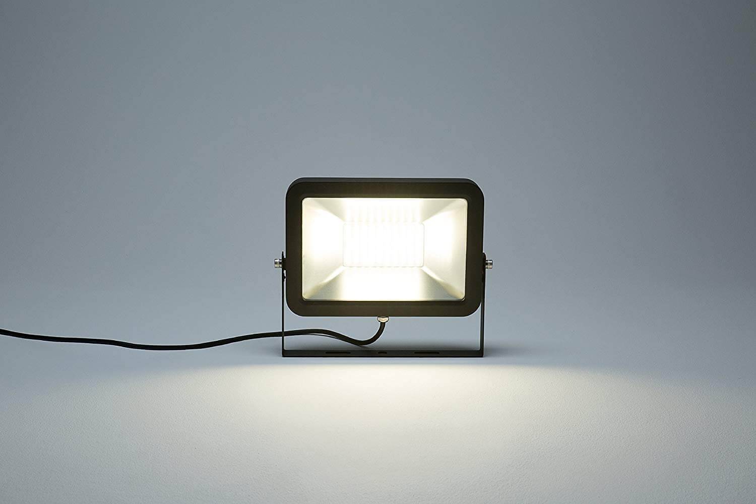 Schwarzer rechteckiger LED-Baustrahler auf einem Stativ mit Stromkabel, der gegen einen einfachen grauen Hintergrund hell leuchtet.
