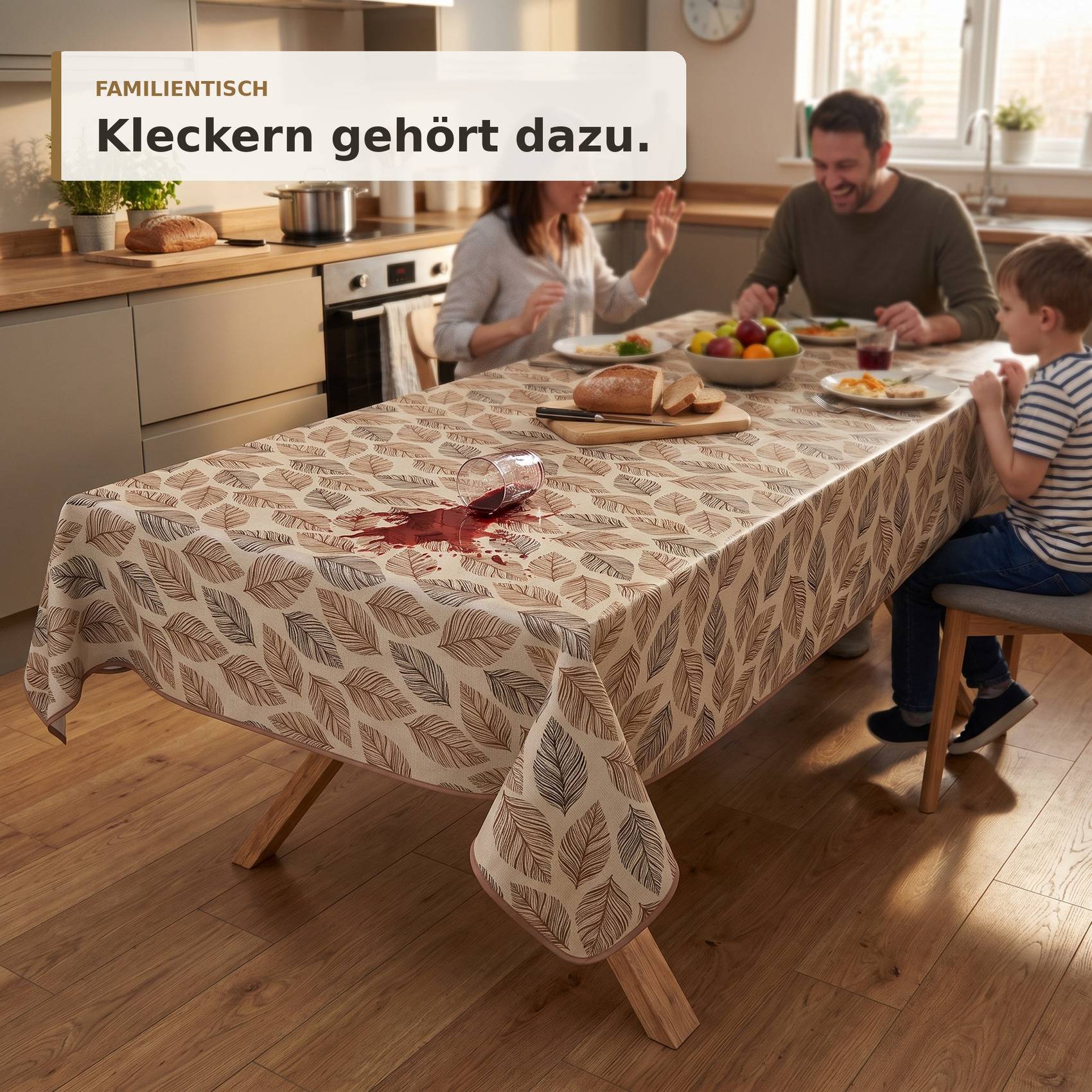 Familienszene in einer Küche mit einem gemusterten Tischtuch, das einen Tisch nach einem Rotweinfleck schützt; Text lautet „FAMILIENTISCH Kleckern gehört dazu."