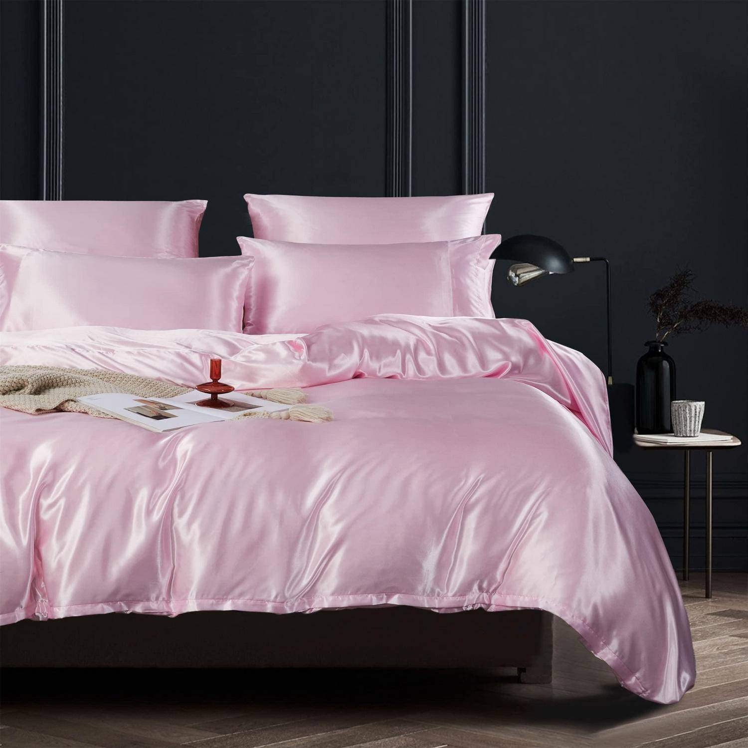 Bett mit glänzender rosa Satinbettwäsche und Kissen, gestylt in einem dunklen modernen Schlafzimmer mit einer Nachttischlampe und kleinem Tisch.