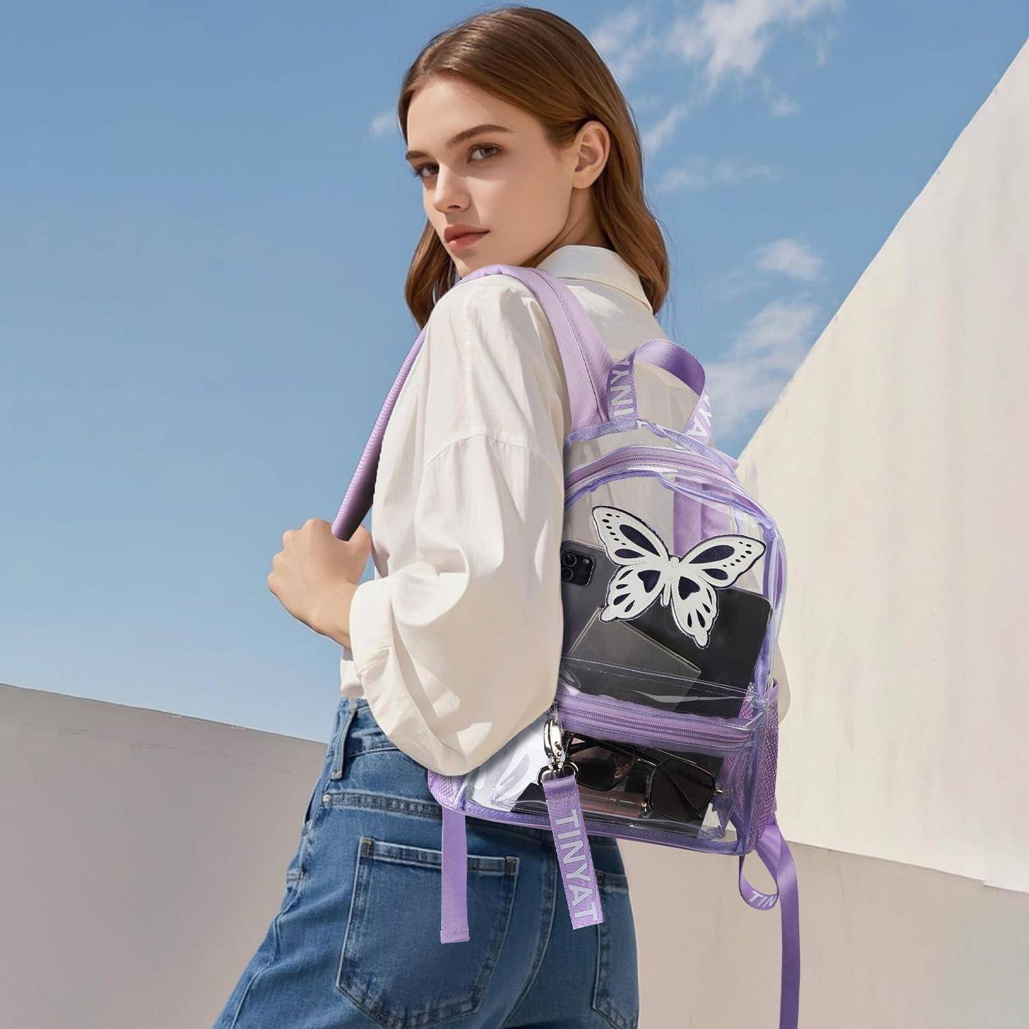 Junge Frau im Freien, die ein weißes Hemd und Jeans trägt und einen transparenten lavendelfarbenen Rucksack mit einer Schmetterlingsdekorationen gegen einen blauen Himmel und eine weiße Wand trägt.