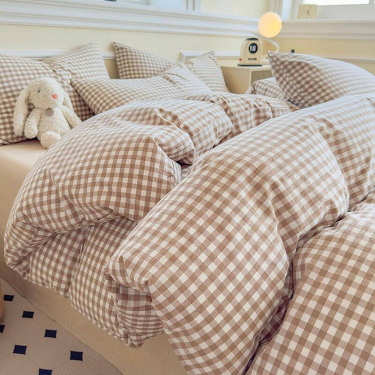 Ein gemütliches Bett mit beigefarbener Gingham-Bettwäsche und passenden Kissen, plus ein kleines Plüschkaninchen auf der linken Seite.