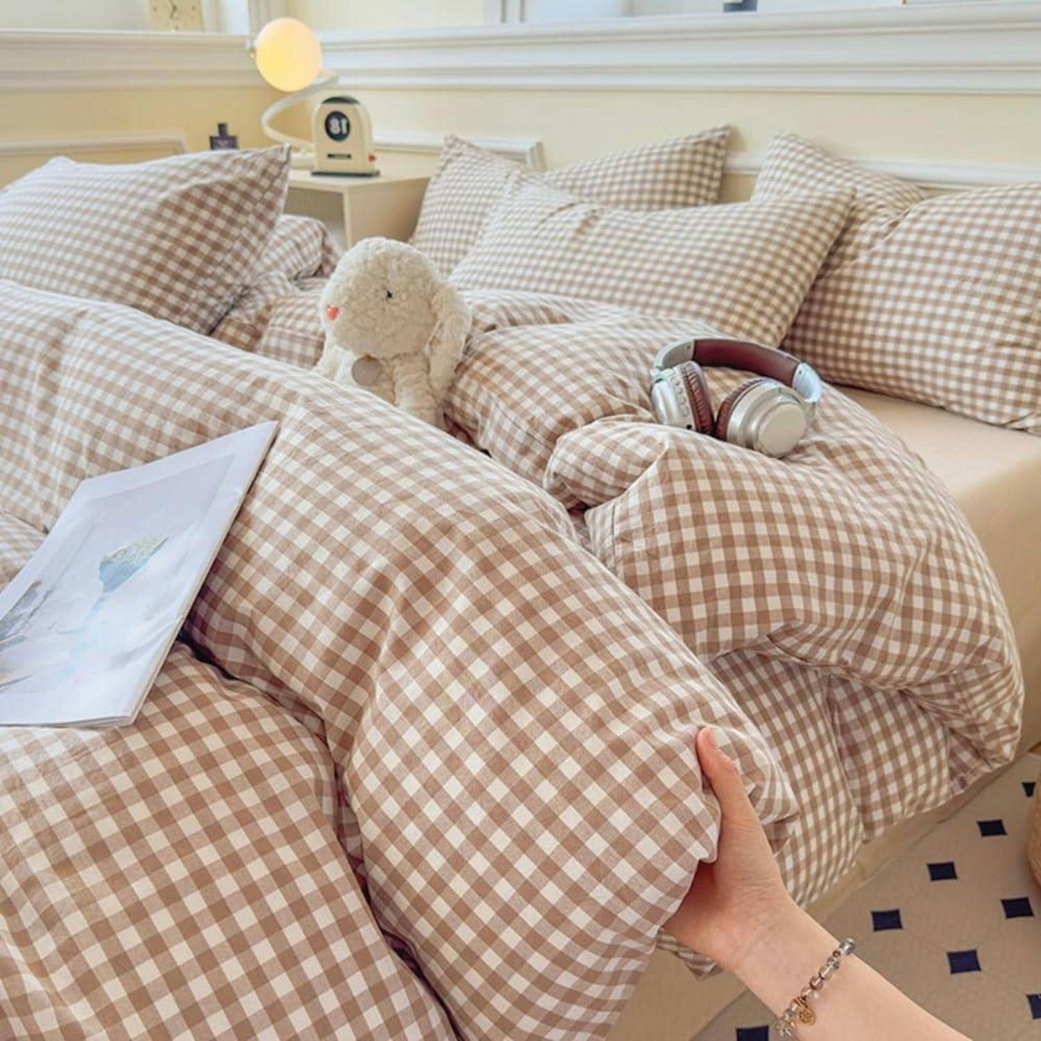 Eine Hand, die ein Gingham-Kissen auf einem Bett hält, das mit passenden Kissen, einem Teddybär, Kopfhörern und einem Buch in einem gemütlichen Schlafzimmer bedeckt ist.