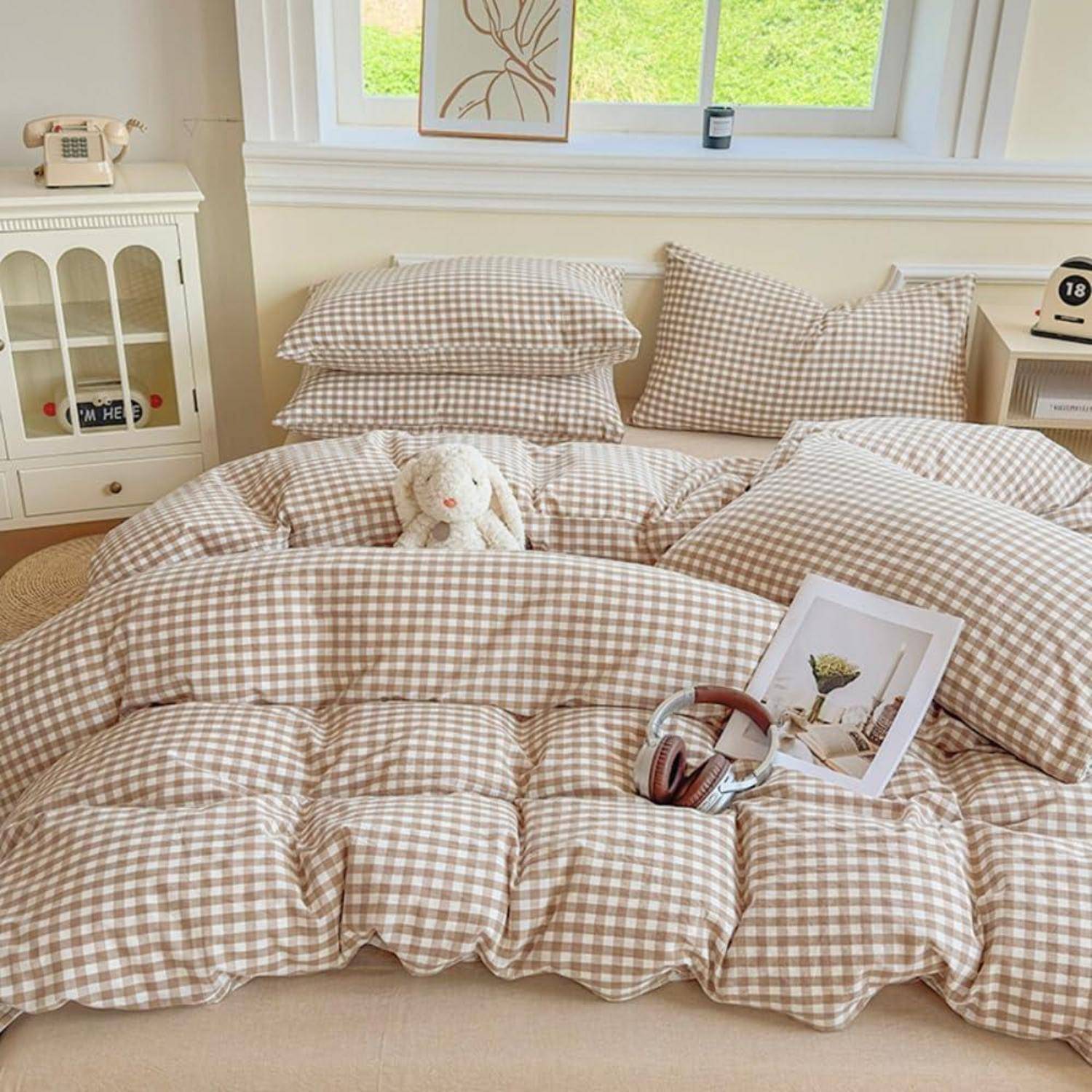 Ein ordentlich gemachtes Bett mit beiger Gingham-Bettwäsche, Kissen, einem Kuschelhase, Kopfhörern und einem Fotodruck in einem hellen Schlafzimmer.
