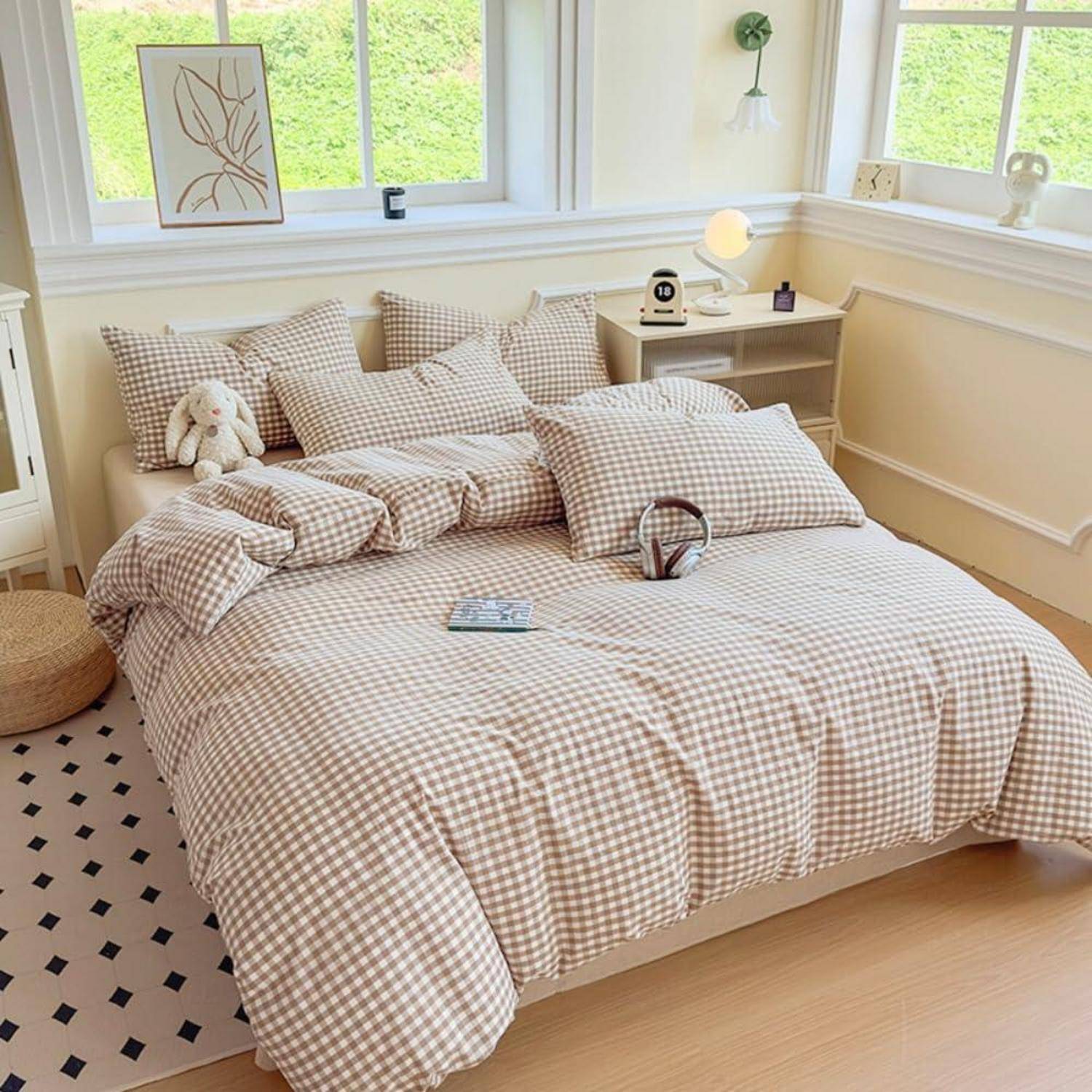 Gemütliches Schlafzimmer mit einem ordentlich gemachten Bett in beigem Gingham-Bettzeug, Kissen, Nachttisch, Lampe und großen Fenstern, die den Raum mit Tageslicht füllen.