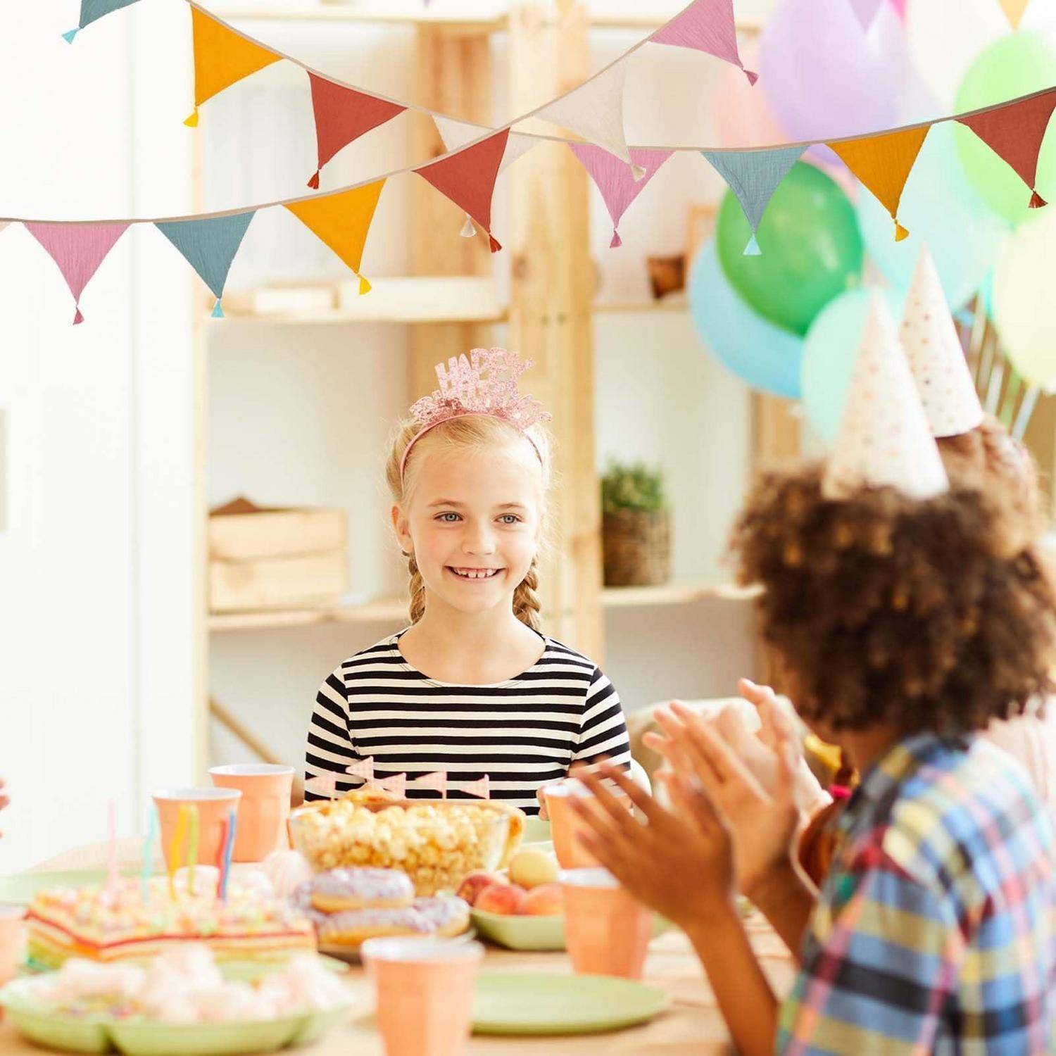 Kinder bei einer Geburtstagsfeier versammeln sich um einen Tisch mit Snacks und Pastellbechern; ein lächelndes Mädchen trägt eine Tiara und Partydekoration hängt über ihnen.