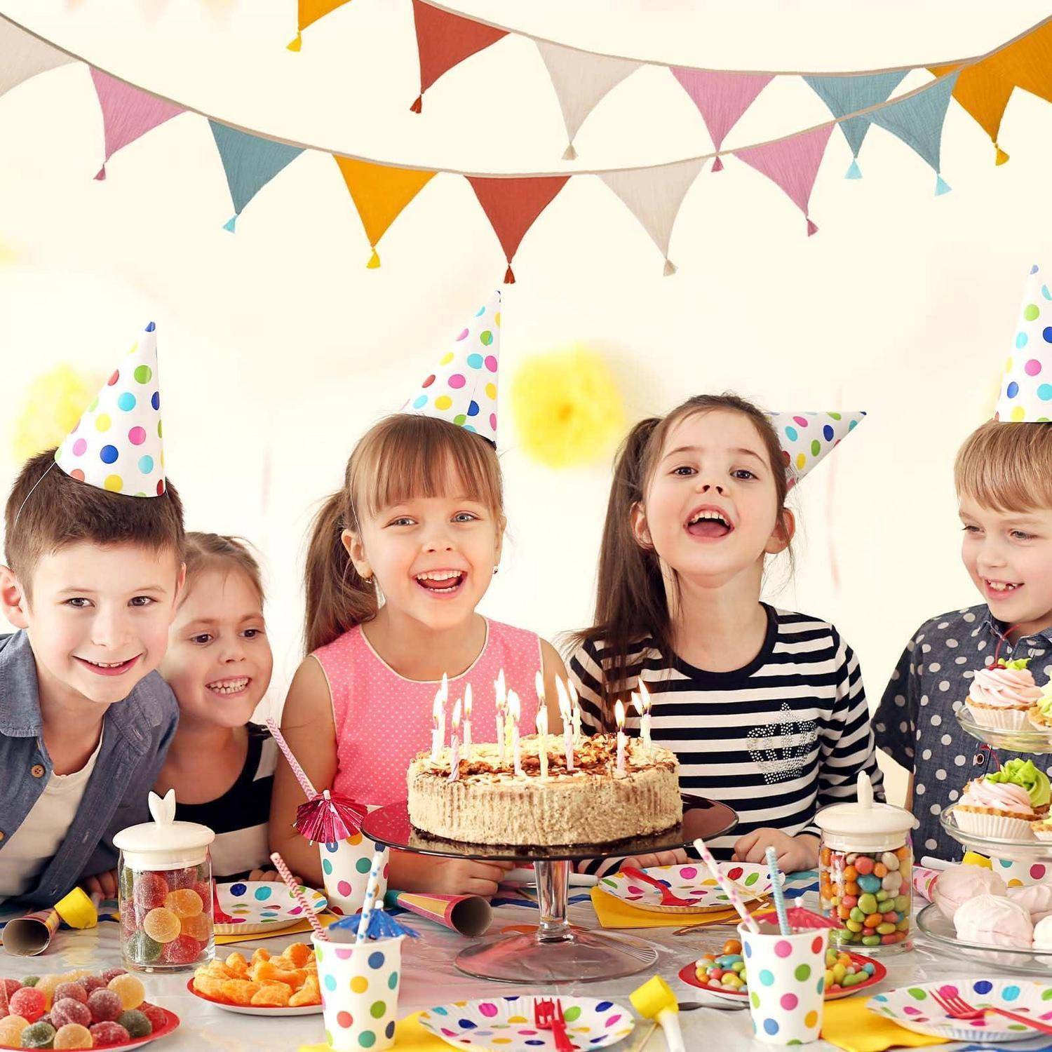Kinder, die Partyhüte tragen, lächeln um einen Geburtstagskuchen mit brennenden Kerzen, Süßigkeiten und bunten Dekorationen an einem Partytisch.