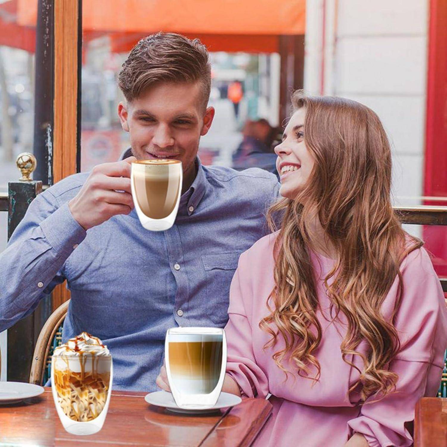 Lächelndes Paar in einem Café, wobei der Mann eine Kaffeetasse hält und die Frau ihn anschaut; zwei weitere Kaffeegetränke stehen auf dem Tisch.