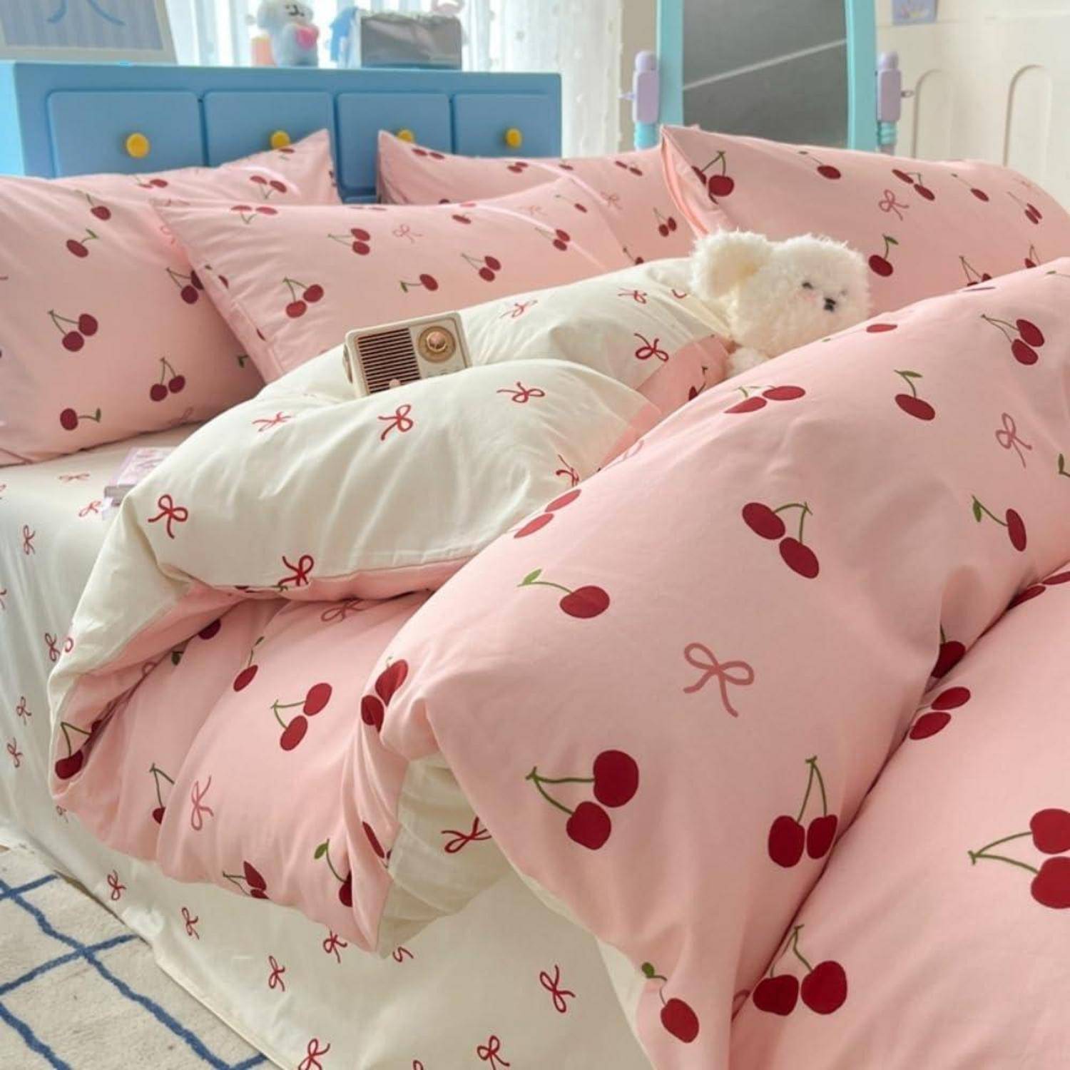 Ein kleiner weißer Hund schaut aus rosa kirschengemusterter Bettwäsche auf einem Bett in einem hellen Schlafzimmer hervor.