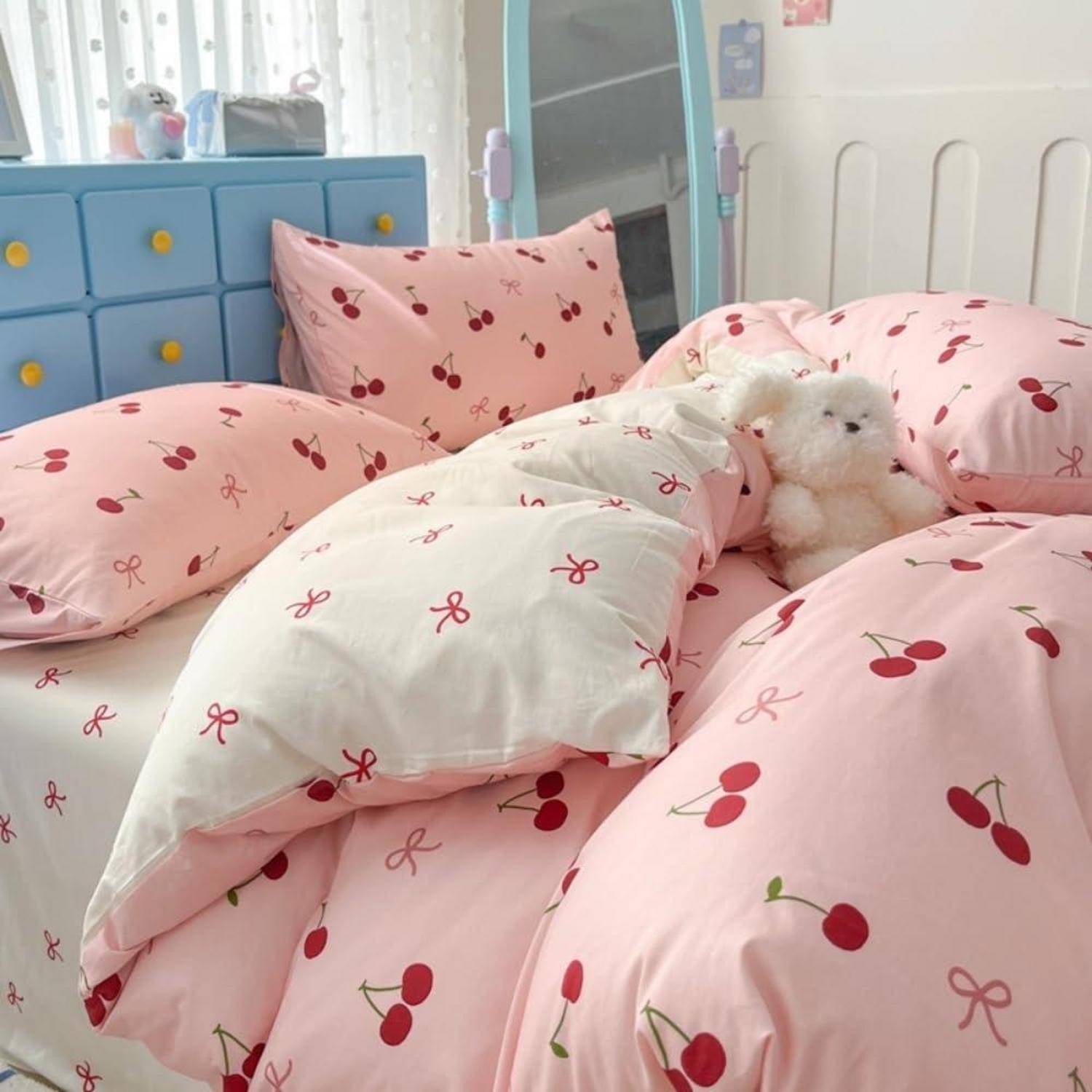 Ein Bett mit rosa Kirschmuster-Bettwäsche und Kissen, mit einem weißen Kuschelbär, der gemütlich zwischen den Decken in einem Schlafzimmer verschachtelt ist.