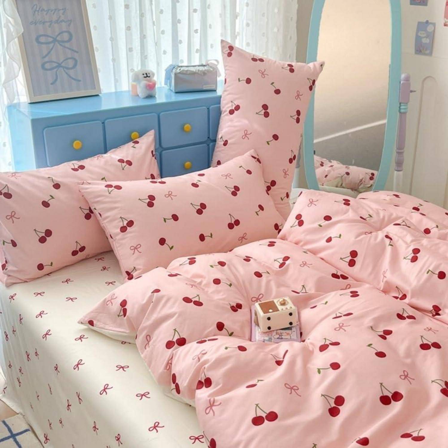 Ein ordentlich gemachtes Bett mit rosa Kirschmuster-Bettwäsche und Kissen in einem Pastellschlafzimmer mit einer blauen Kommode und weichen Vorhängen.