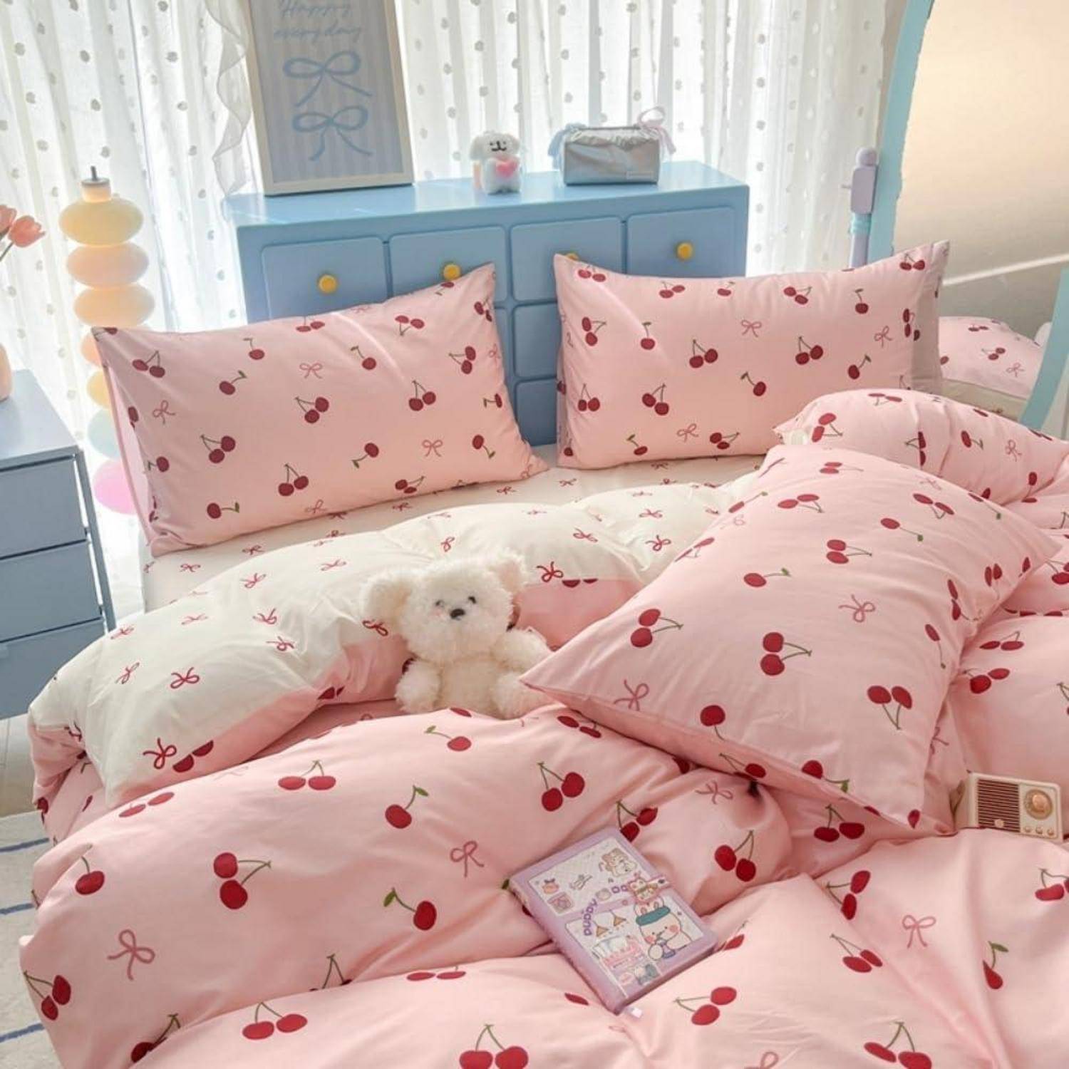 Ein ordentlich gemachtes Bett mit rosa Kirschdruck-Bettwäsche und Kissen, ein kleiner Teddybär und eine pastellblaue Kommode in einem hellen Schlafzimmer.