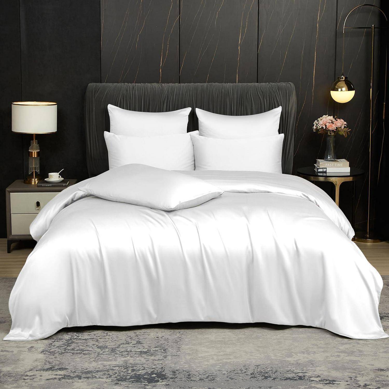 Ein ordentlich gemachtes Bett mit weißen Kissen und seidenartig weißer Bettwäsche in einem dunklen modernen Schlafzimmer, flankiert von Nachttischlampen und Dekor.