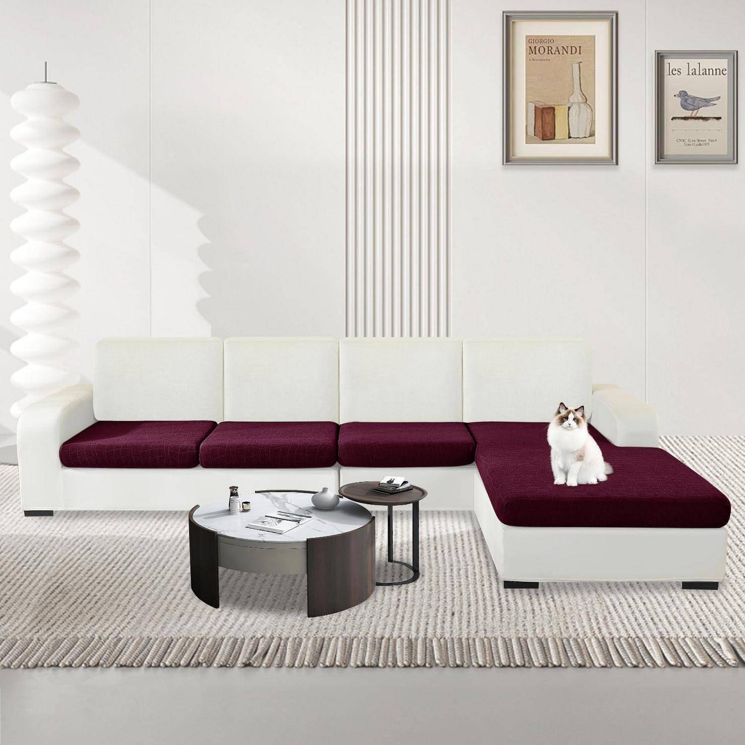Modernes weißes Ecksofa mit bordeauxfarbenen Kissen in einem minimalistischen Wohnzimmer, mit einer Katze, die auf der Chaiselongue sitzt, und rahmenkunst an der Wand.