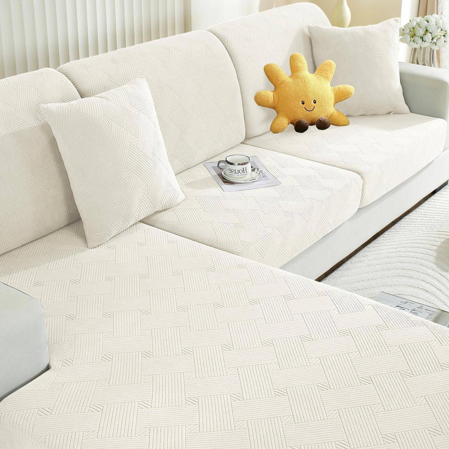 Weißes Sektionalsofa mit strukturierten Bezügen, zwei weiße Kissen, ein sonnenförmiges Plüschtier und eine Tasse auf einem Tablett in einem hellen Wohnzimmer.