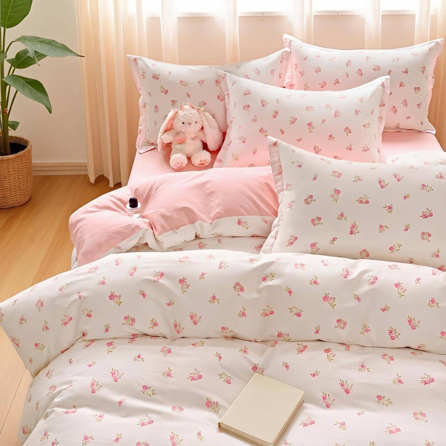 Rosa Blumenbettwäsche mit mehreren Kissen, einem kuscheligen Hase und einem kleinen Notizbuch auf einem Bett in einem hellen Schlafzimmer.