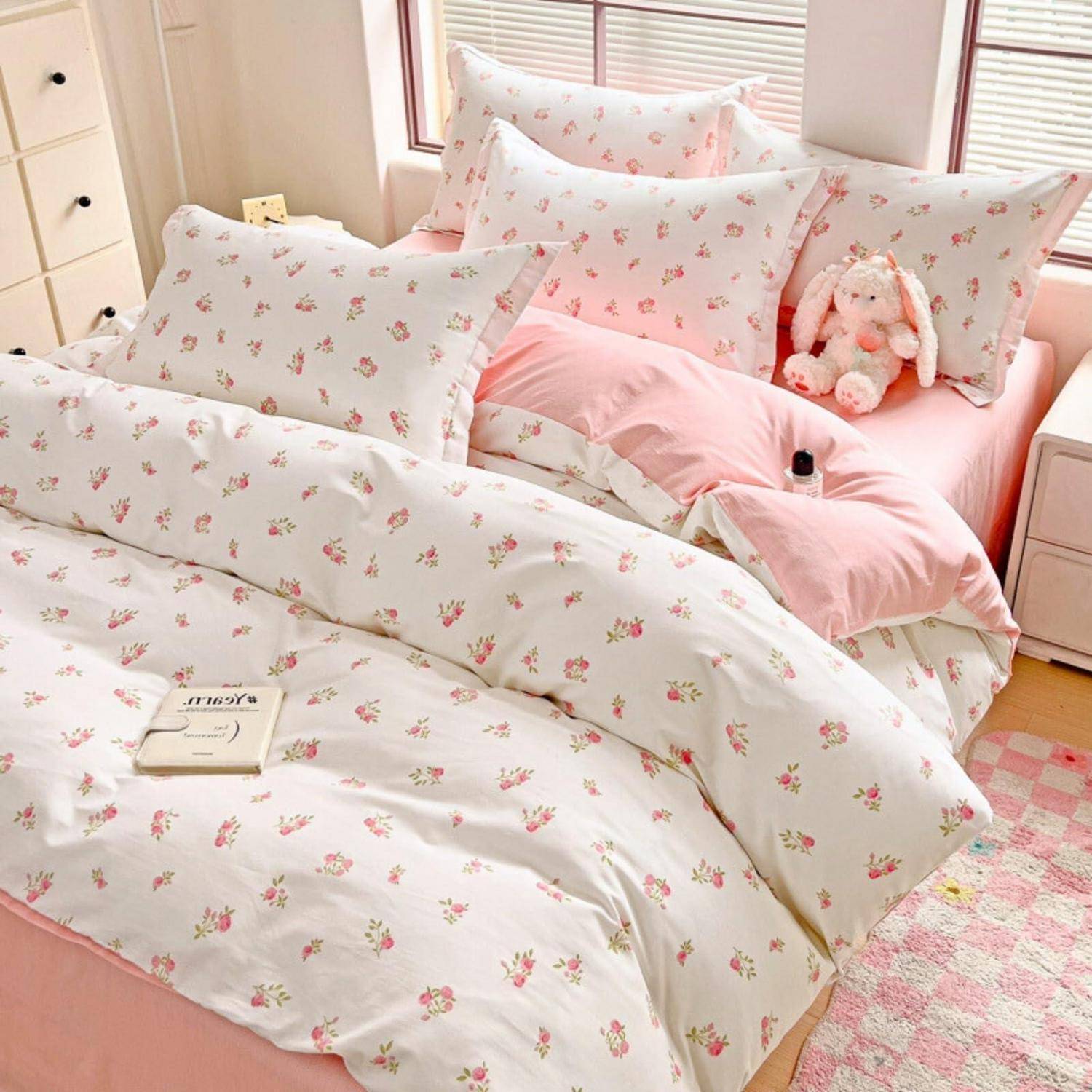 Rosa Blumenbettwäsche auf einem ordentlich gemachten Bett mit Kissen, einem Plüschhäschen und einem kleinen Buch in einem hellen Schlafzimmer.
