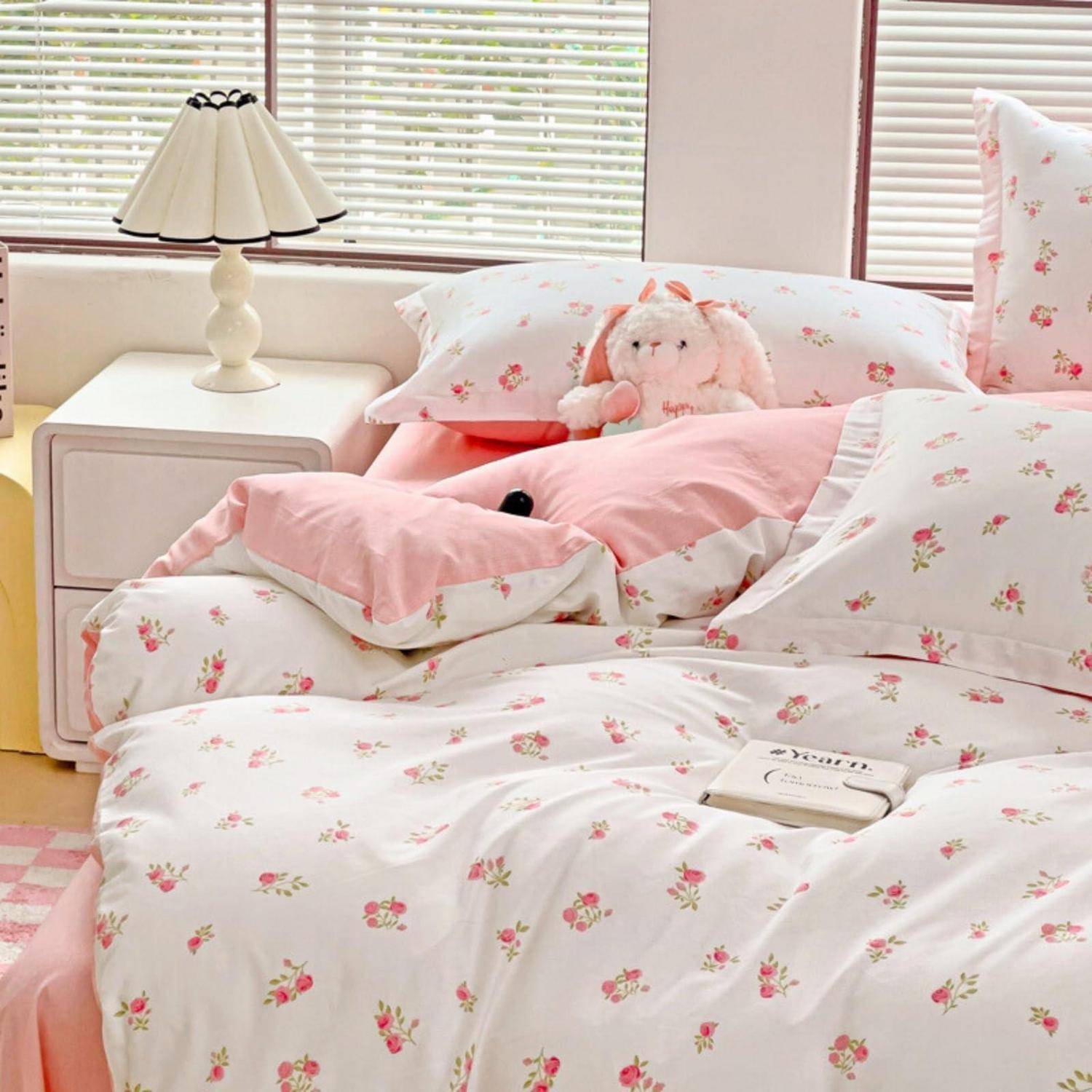 Rosafarbene Blumenbettwäsche auf einem ordentlich gemachten Bett mit Kissen und einem Plüschhäschen, neben einem weißen Nachttisch und einer Lampe bei einem Fenster.