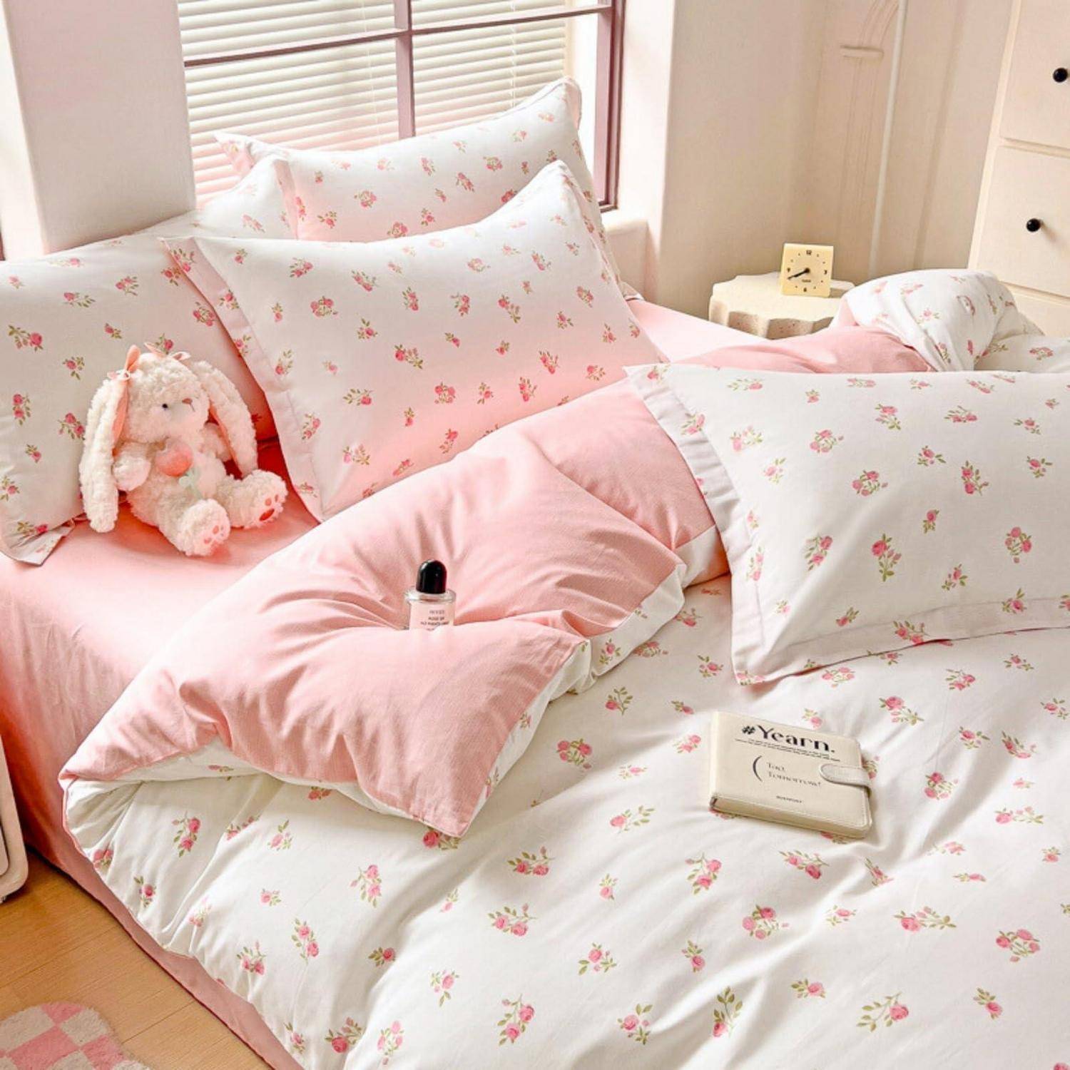 Rosa Blumenbettwäsche auf einem ordentlich gemachten Bett mit Kissen, einem Plüschhäschen, einer kleinen Flasche und einem Buch in einem gemütlichen, sonnendurchfluteten Schlafzimmer.