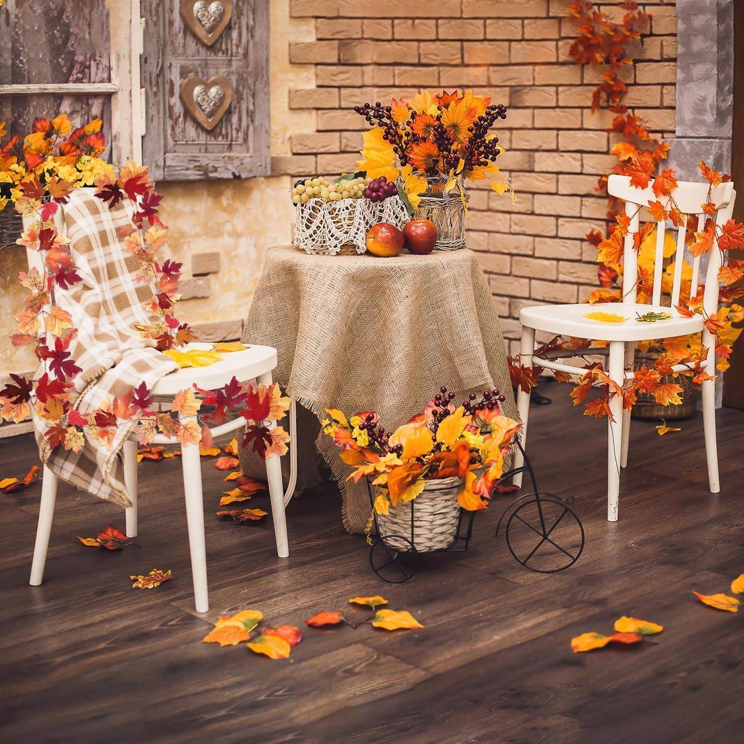 Herbstlich dekorierte Terrasse mit zwei weißen Stühlen, einem mit Sackleinen bezogenen Tisch und Herbstblättern, Kürbissen und Blumen, die die Szene schmücken.