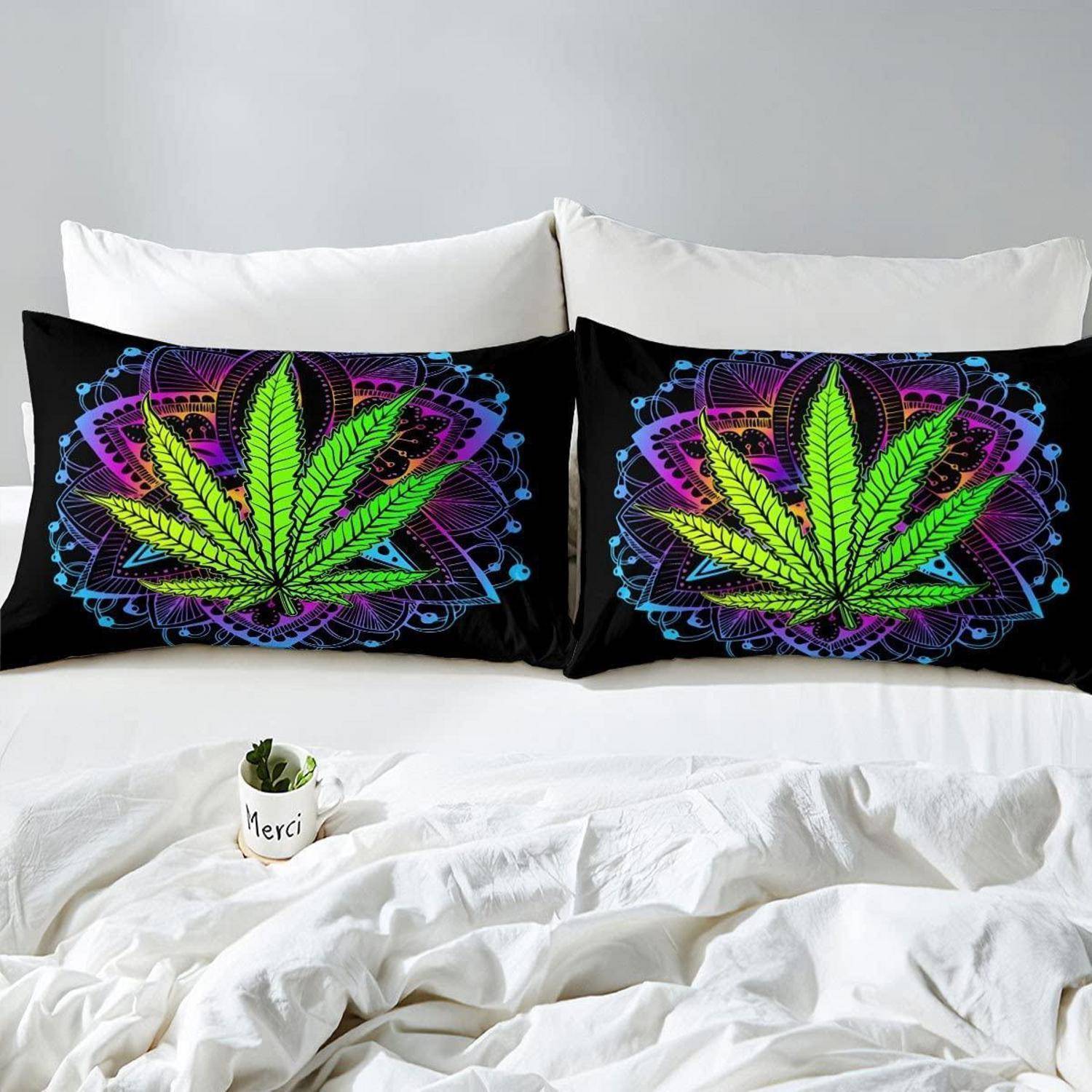 Bett mit zwei schwarzen dekorativen Kissen mit Neon-Stil-Cannabisblatt-Designs, weißer Bettwäsche und einer kleinen "Merci""-Tasse mit einer Pflanze.