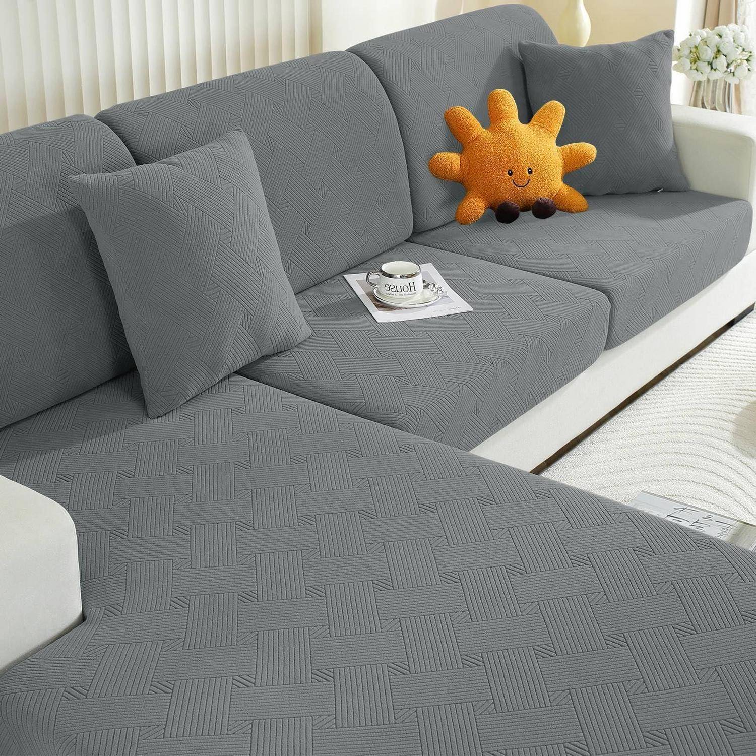 Graues L-förmiges Sofa mit strukturiertem Sofabezug und Kissen, auf dem eine Kaffeetasse auf einer Zeitschrift und ein orangefarbenes lächelndes Plüschtier stehen.