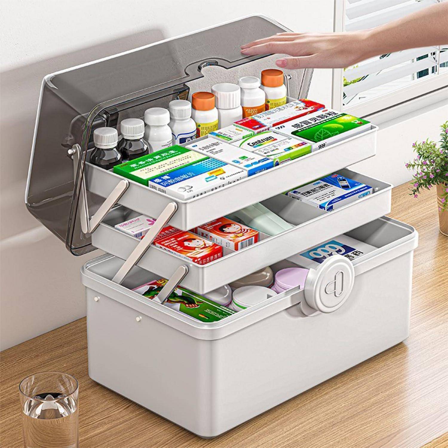 Hand öffnet eine gestaffelte weiße Medikamentenaufbewahrungsbox mit ausziehbaren Tabletts, die Rezeptflaschen, Salben und Erste-Hilfe-Artikel enthalten.