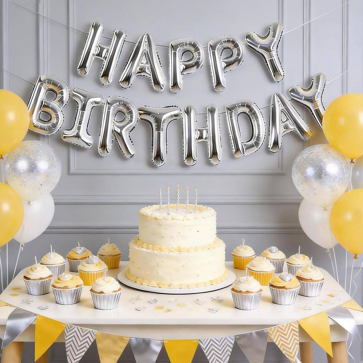 Geburtstagsparty-Setup mit silbernen „HAPPY BIRTHDAY"-Luftballons, einer zweistöckigen Torte mit Kerzen, Cupcakes und gelb-weißen Luftballons und Wimpelketten.