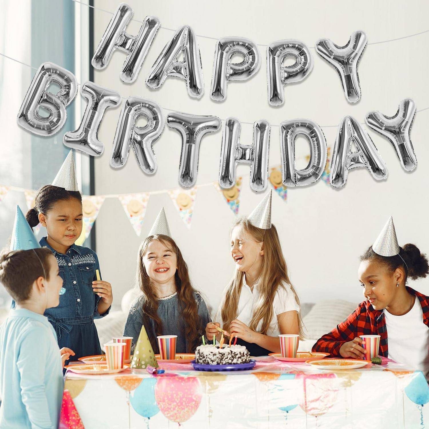 Kinder mit Partyhüten feiern einen Geburtstag um einen dekorierten Tisch mit Kuchen, Snacks und „HAPPY BIRTHDAY"-Luftballons.