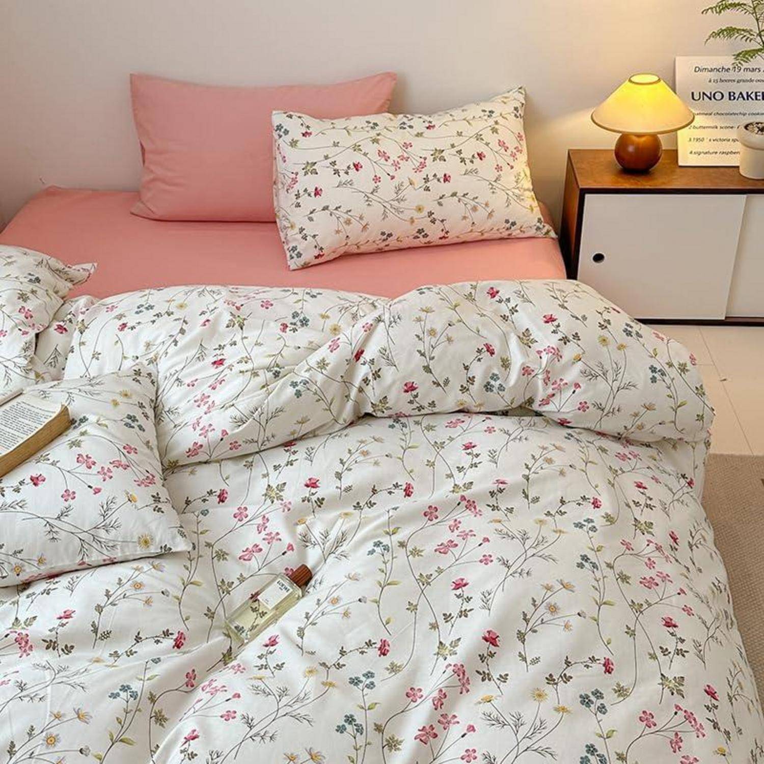 Rosa Bett mit floralem Bettdecke und Kissen neben einer kleinen Lampe auf einem Nachttisch in einem gemütlichen Schlafzimmer.