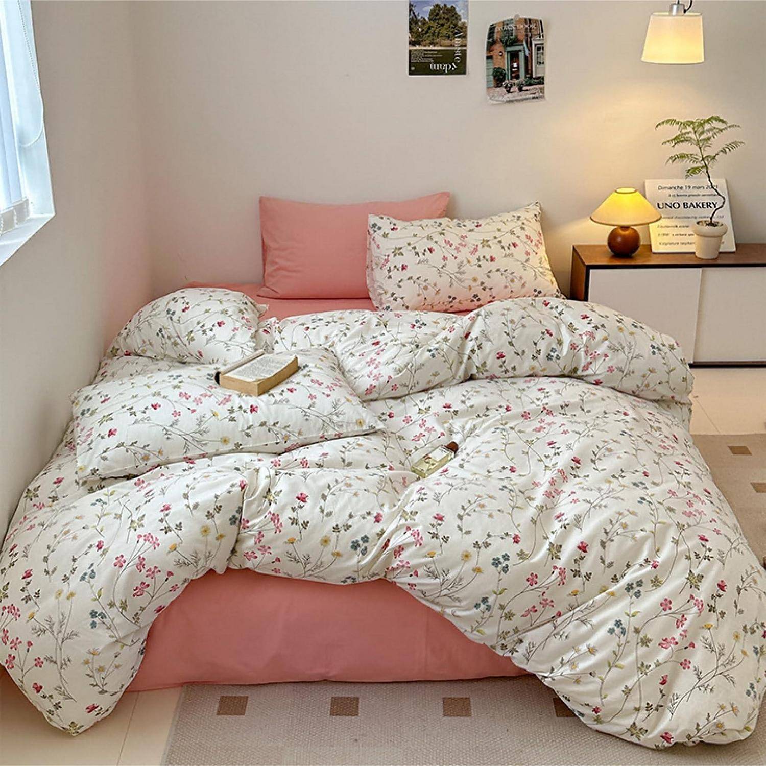 Gemütliches Schlafzimmer mit unmachtem Blumenbettzeug, rosa Kissen und Bettdecke, ein Buch auf dem Bett und eine Nachttischlampe auf einem kleinen Tisch.