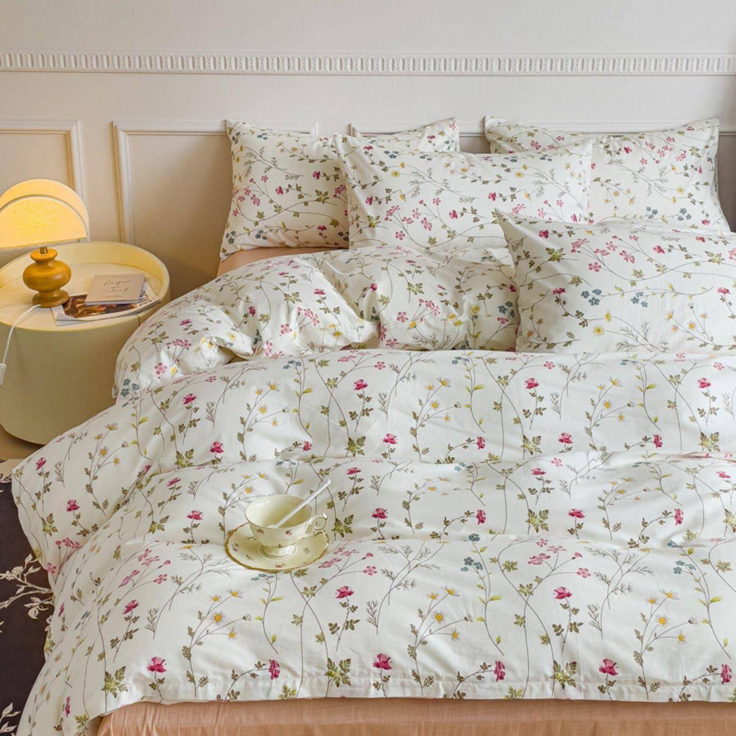 Ein ordentlich gemachtes Bett mit floralem Bettzeug und Kissen, neben einem runden Nachttisch mit einer brennenden Lampe und einem Buch in einem sanft beleuchteten Schlafzimmer.