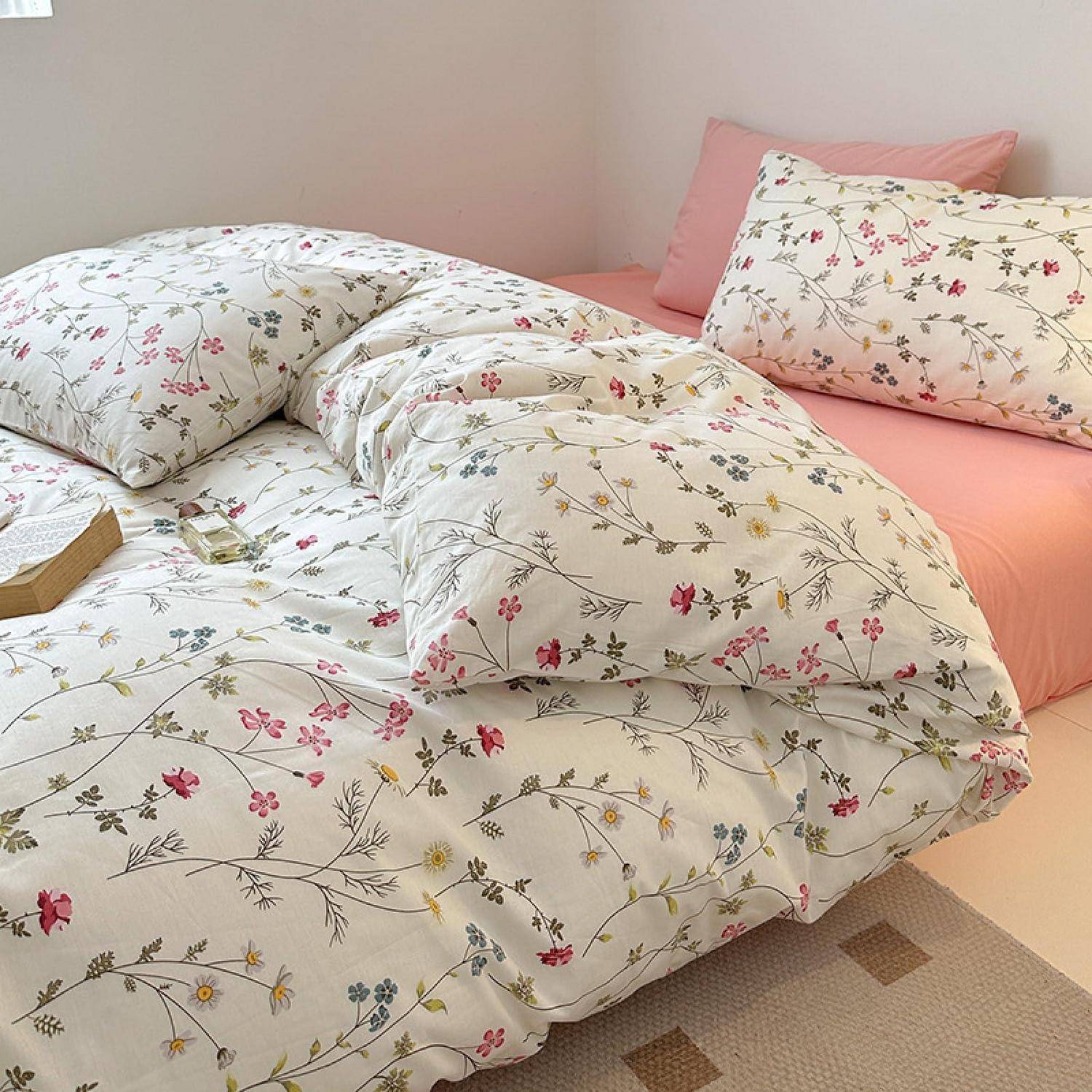 Ein ordentlich gemachtes Bett mit weiß gemusterter Bettwäsche und Kissen mit Blumenmotiven, akzentuiert durch ein rosa Spannbettuch und ein Kissen vor einer hellfarbigen Wand.