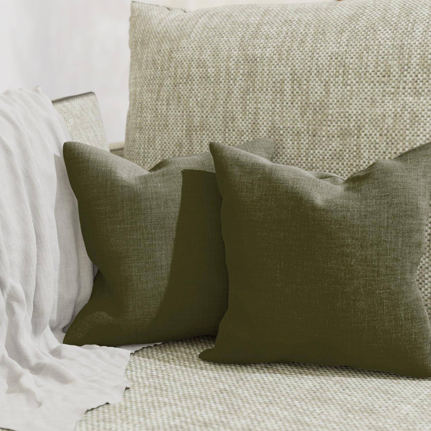 Zwei olivgrüne Dekokissen auf einem beigen strukturierten Sofa neben einer drappierten weißen Decke.
