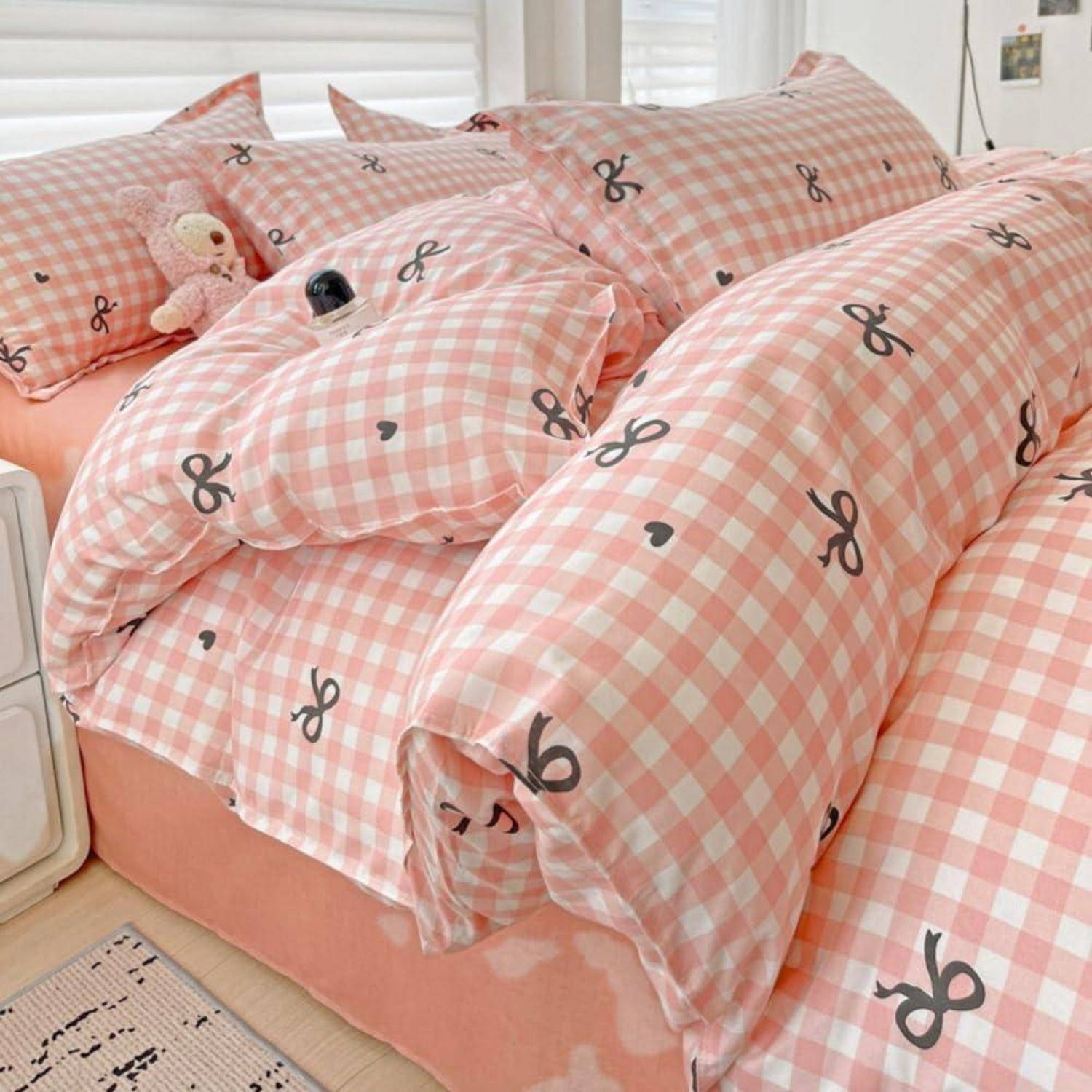 Rosa Gingham-Bettwäsche mit Schleifendruck und einer passenden Decke auf einem ordentlich gemachten Bett, mit einem kleinen Plüschtier in der Nähe der Kissen.