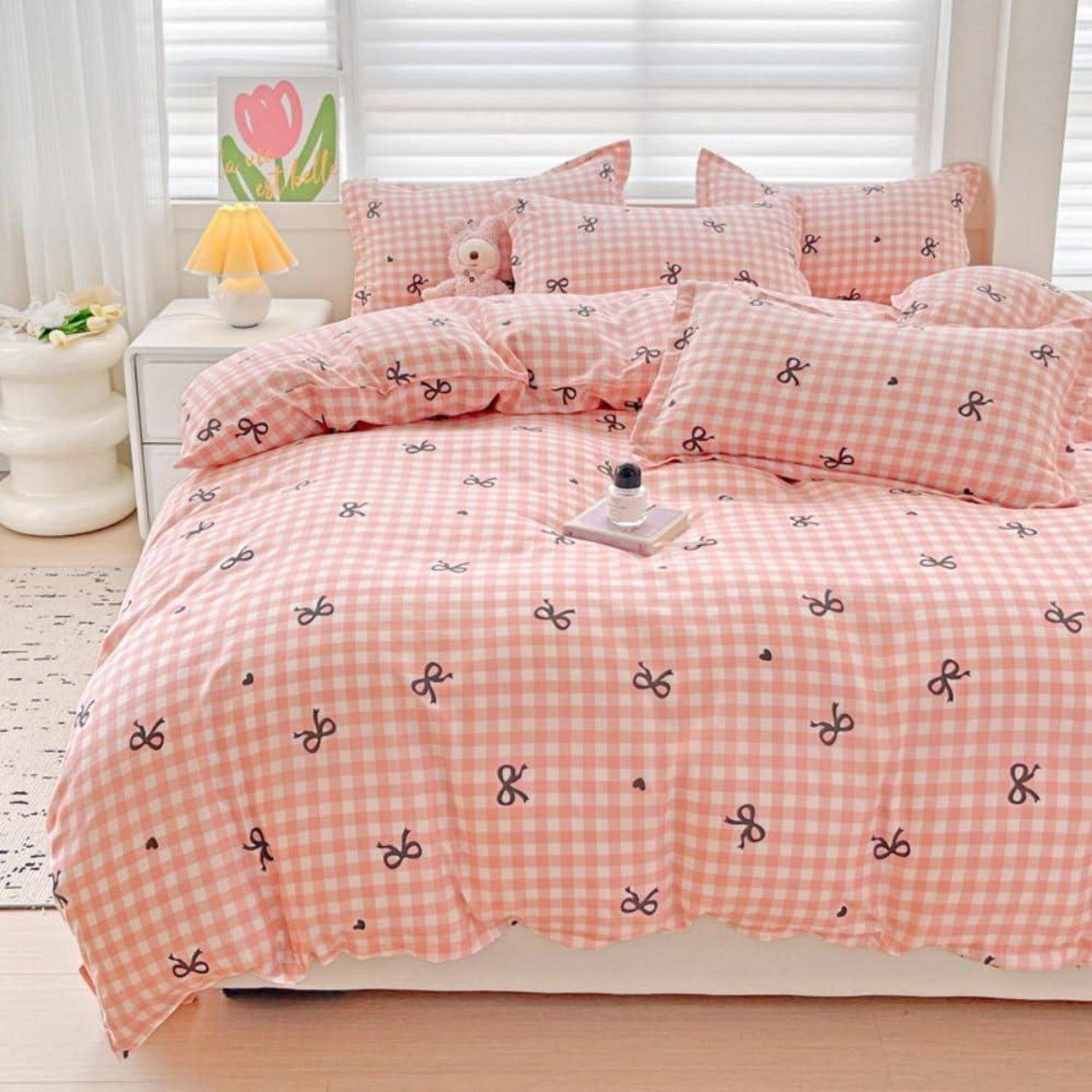 Rosafarbene Gingham-Bettwäsche mit schwarzem Schleifendruck auf einem ordentlich gemachten Bett, in einem hellen Schlafzimmer mit weißen Jalousien und einer Nachttischlampe.