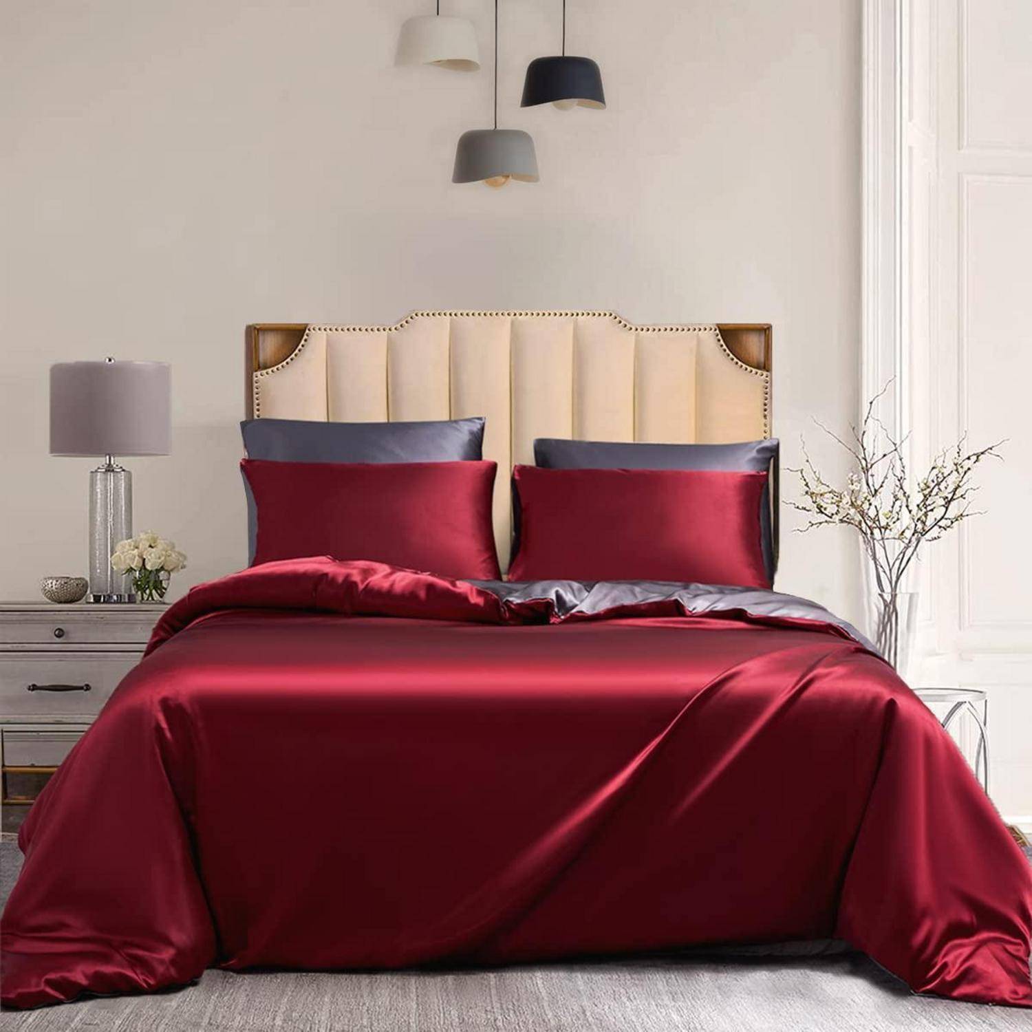 Ein ordentlich gemachtes Bett mit tiefroter Satinbettbezug und Kissen, beige gepolstertem Kopfteil, Nachttischlampe und minimalistischer Schlafzimmerdekoration.