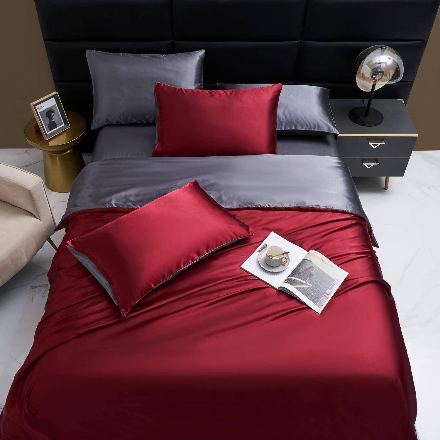 Stilvolles Bett mit burgunderrot und grau Satinbettzeug, rot und grau Kissen und passenden Nachttischen mit Lampe und Dekoration.