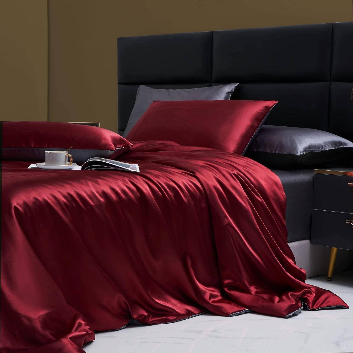 Rote Satinbettwäsche auf einem modernen schwarzen Bett mit Kissen, einer Tasse und einem aufgeschlagenem Buch auf der Decke, in einem minimalistischen Schlafzimmer.