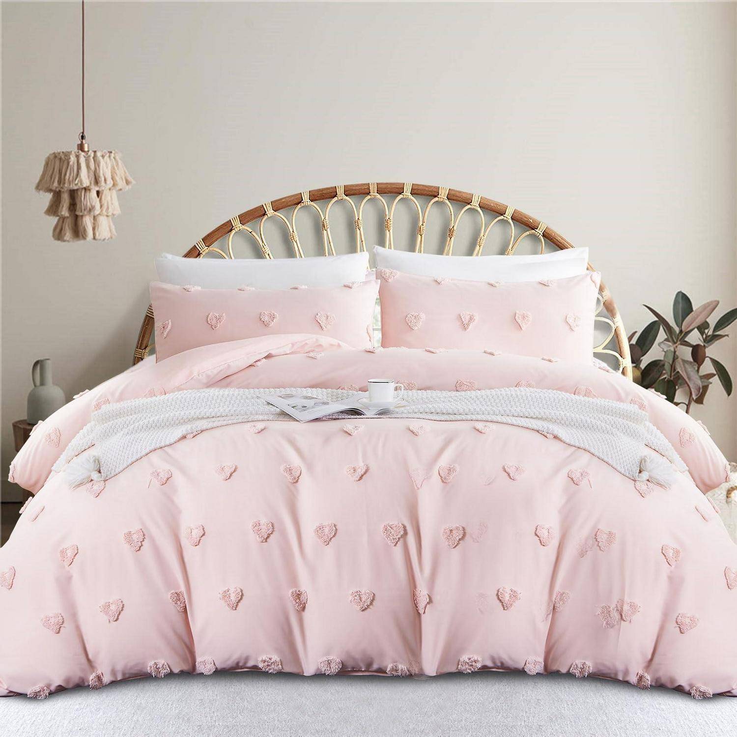 Ein ordentlich gemachtes Bett mit rosa herzgemusterter Bettdecke und Kissen, weißer Decke und einem geflochtenen Rattan-Kopfteil in einem weichen, minimalistischen Schlafzimmer.